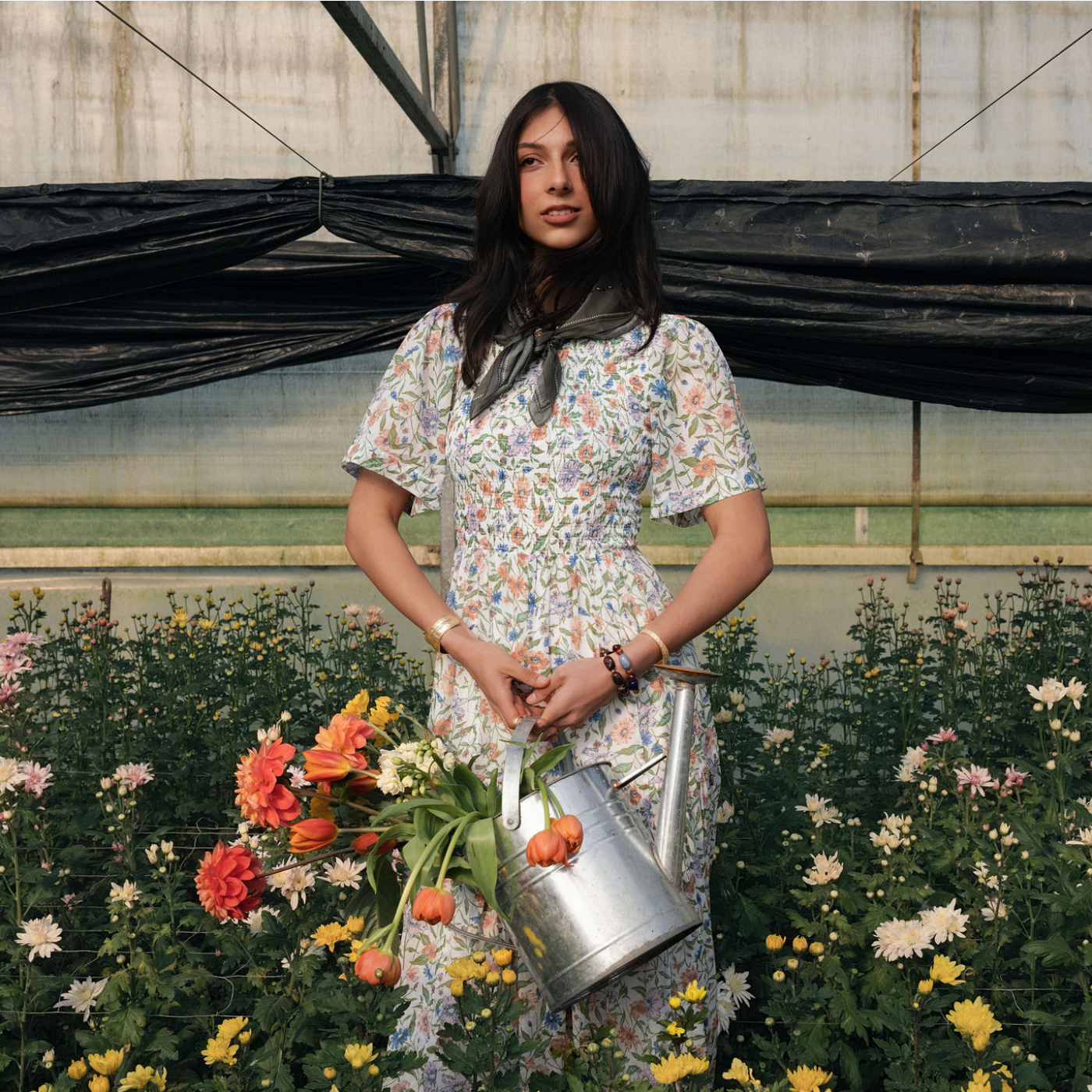 Woman standing in a greenhouse wearing a floral spring dress, holding a watering can filled with flowers.
