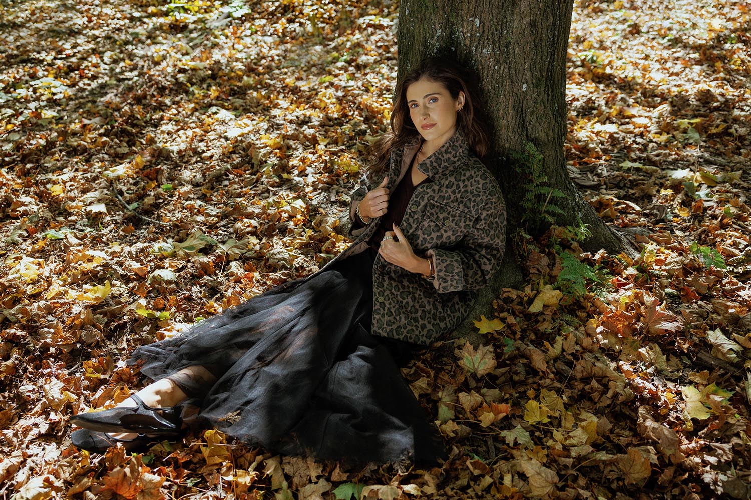 A woman wearing a brown cap, pink sweater, gray quilted jacket, and white pants stands by a river with a forested background, looking to the side.