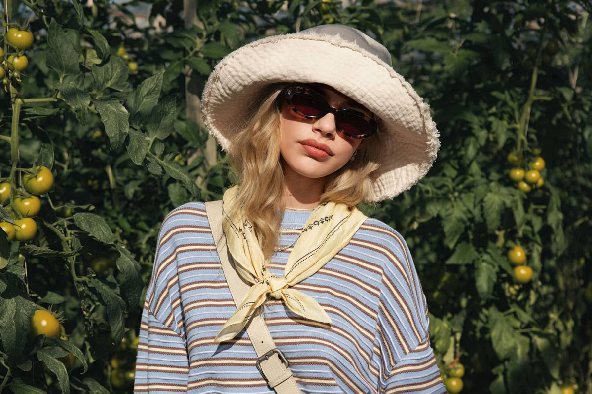A collection of gifts on a blue background, including handbags, a straw hat, sunglasses, mugs, beauty products, a striped shirt, flowers, a candle, playing cards, and a book.