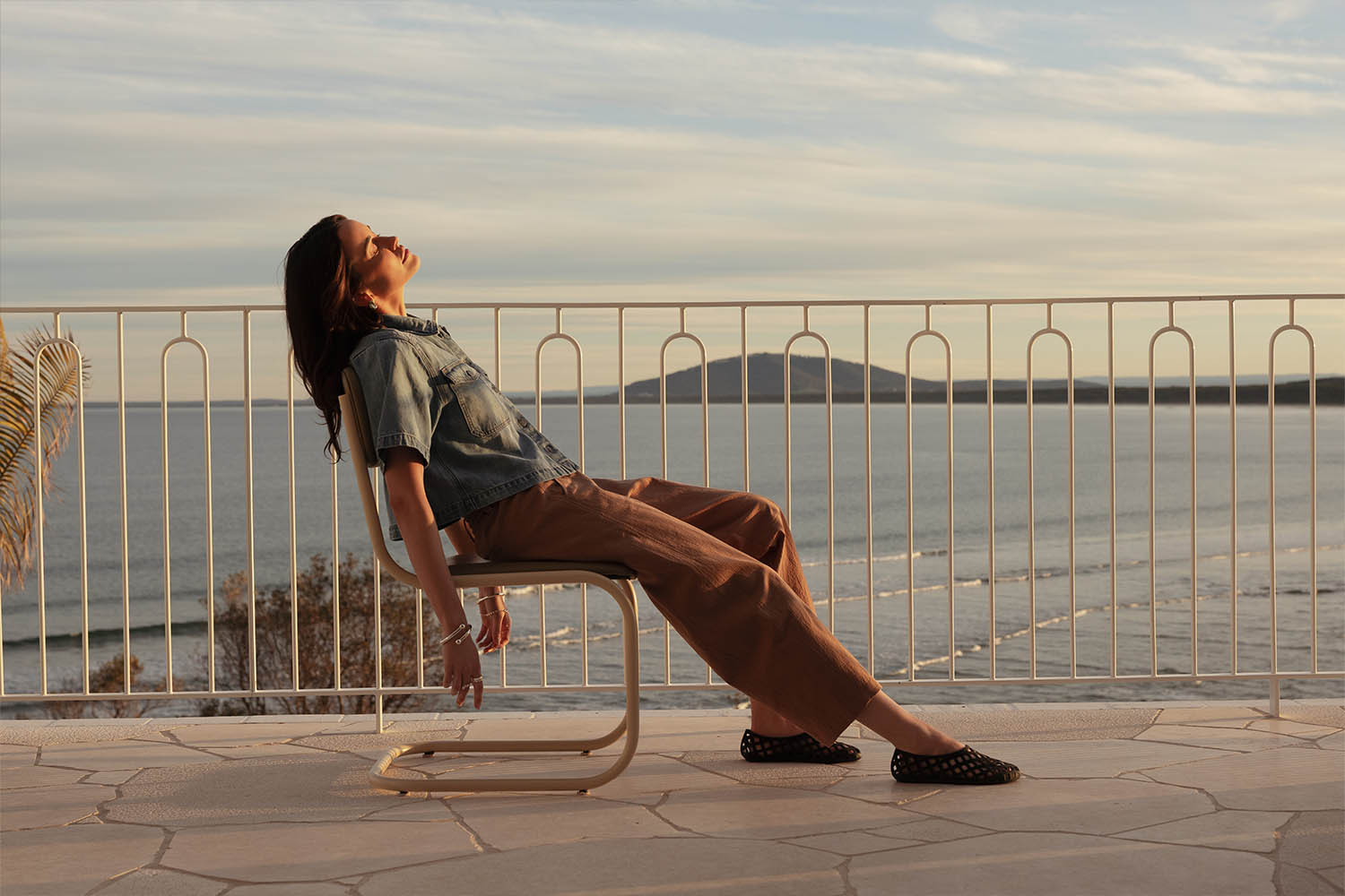 A woman leans back on a chair on a seaside terrace at sunset, eyes closed, enjoying the sunlight, with calm water and distant mountains in the background.
