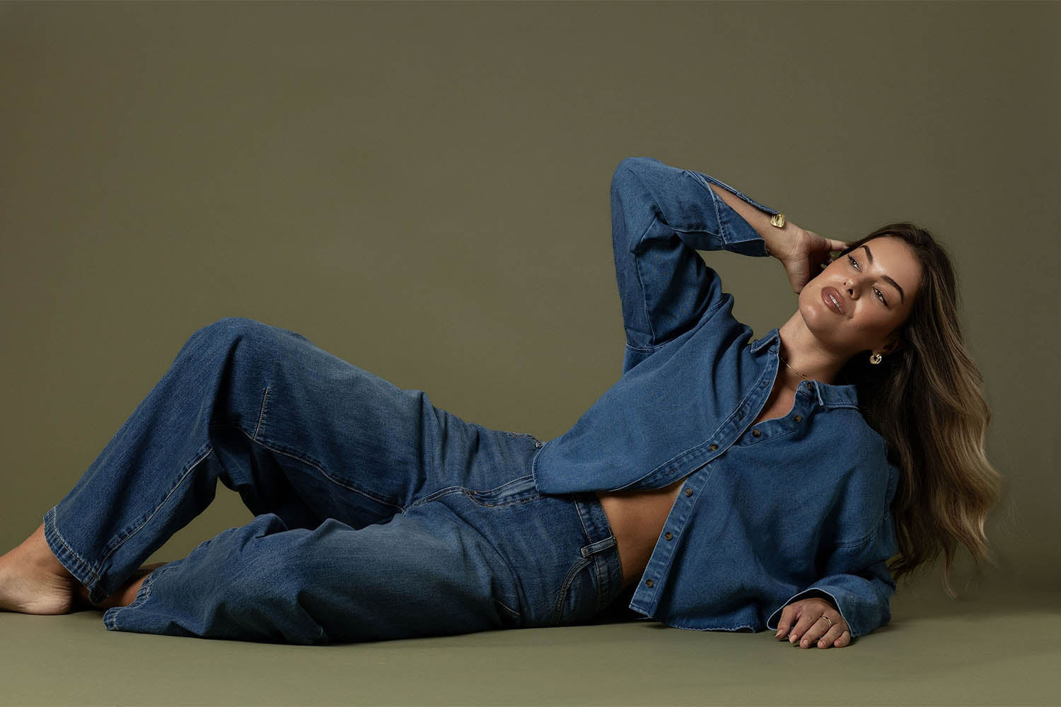 A person wearing a denim jacket and jeans smiles and spreads their arms wide while standing on a sandy beach with waves and blue sky in the background.