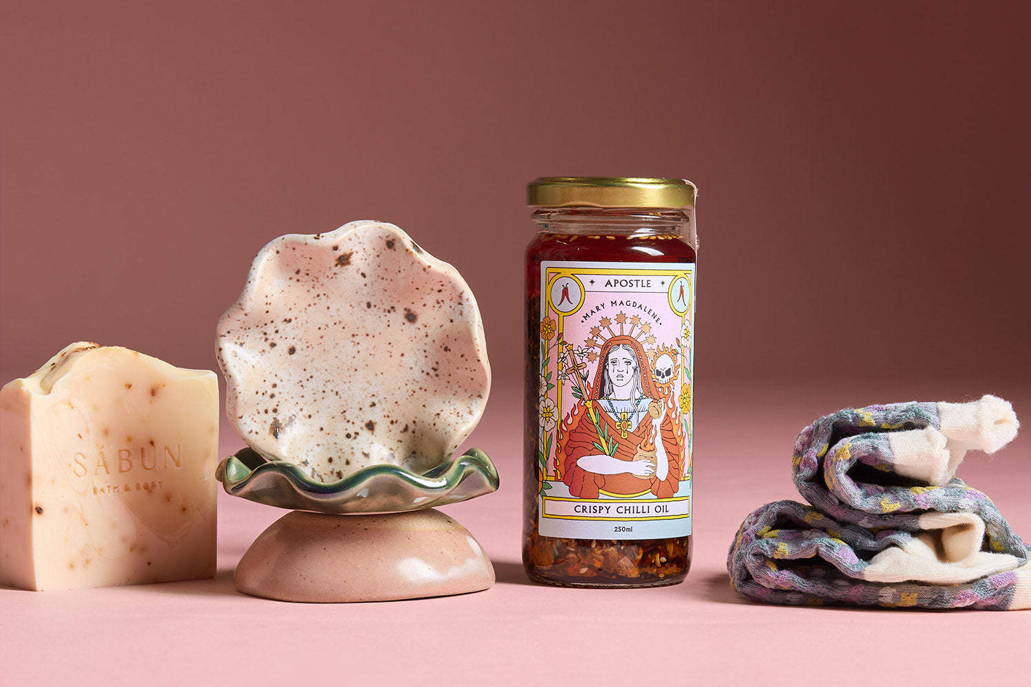 Three jars of Apostle brand seasonings and salts with illustrated labels, stacked in front of a bowl of lemons and pears on a kitchen countertop by a window.