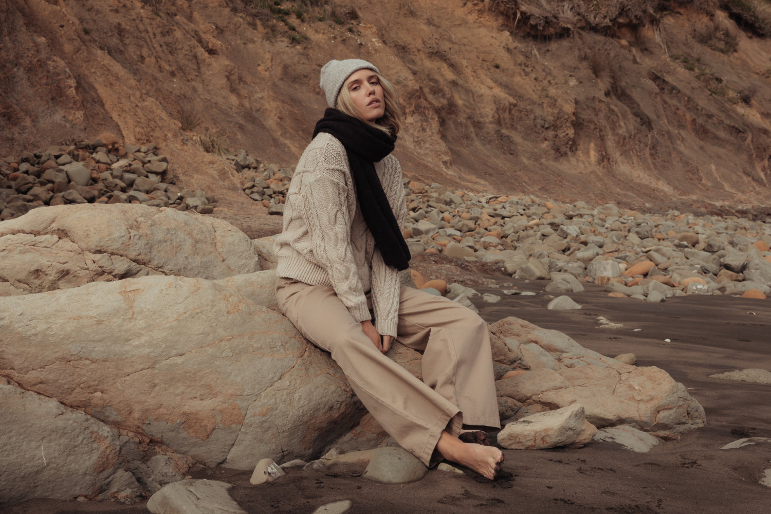 A woman in a printed maxi dress and brown quilted jacket leans on a large rock in a grassy, rocky outdoor landscape under a partly cloudy sky.