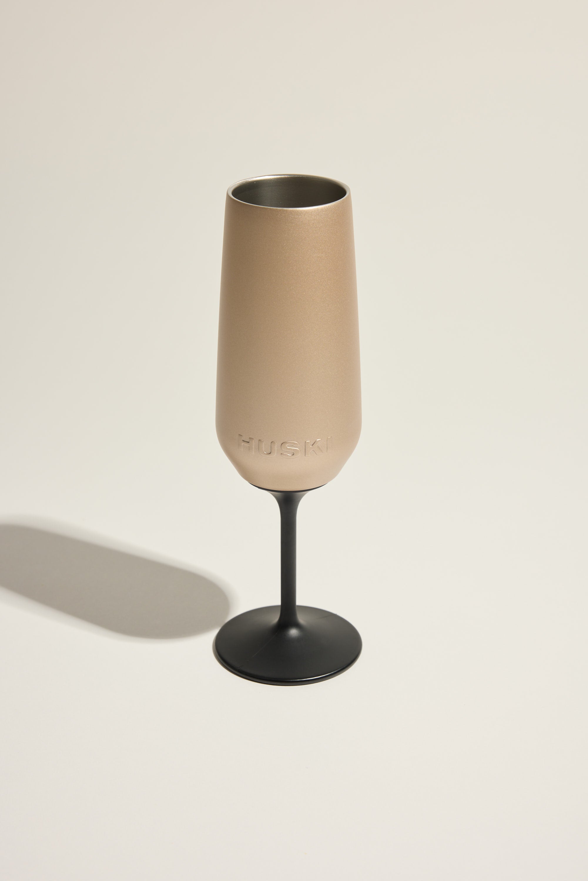 A modern Huski Champagne Flute with insulated matte beige cup, black stem, and embossed HUSKI logo stands on a neutral background, casting a soft shadow—showcasing the premium design from Huski.