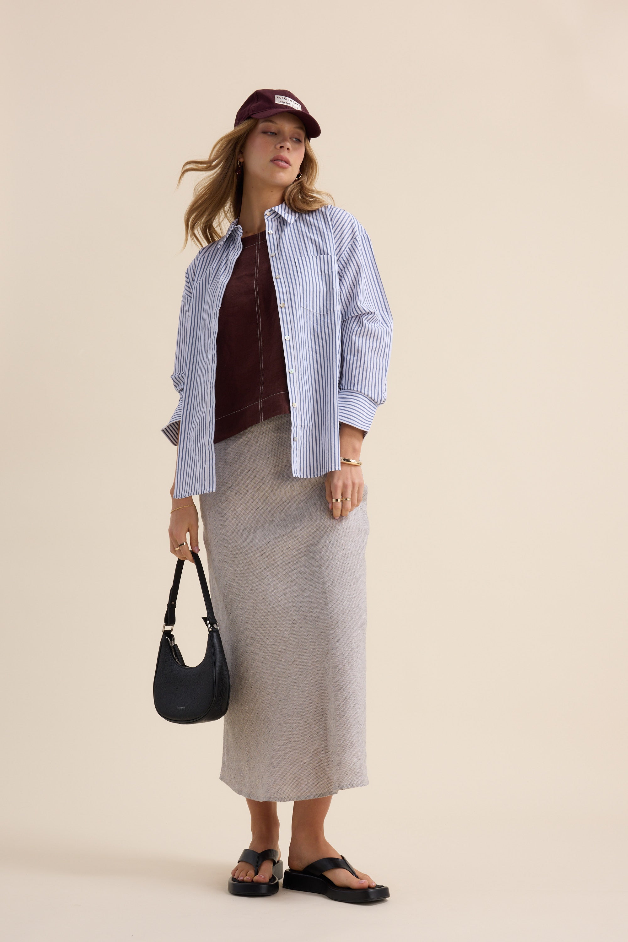 Woman in striped shirt, cap, sandals, Max The Linen Slip Skirt, with black handbag.