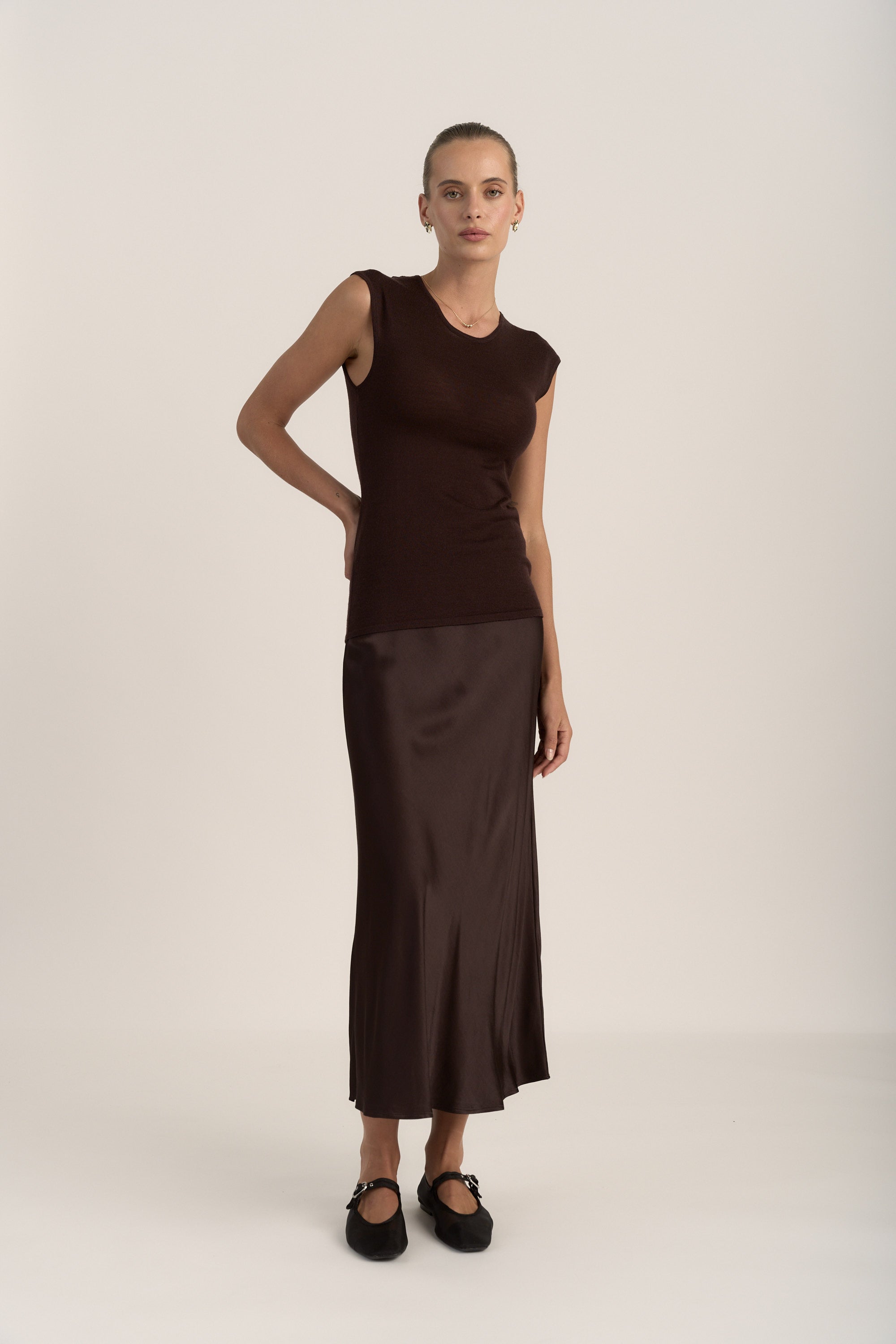 A woman stands against a plain light background wearing Max’s The Slip Skirt in rich brown with a sleeveless dark brown top and black flats, her hair pulled back and one hand on her hip.