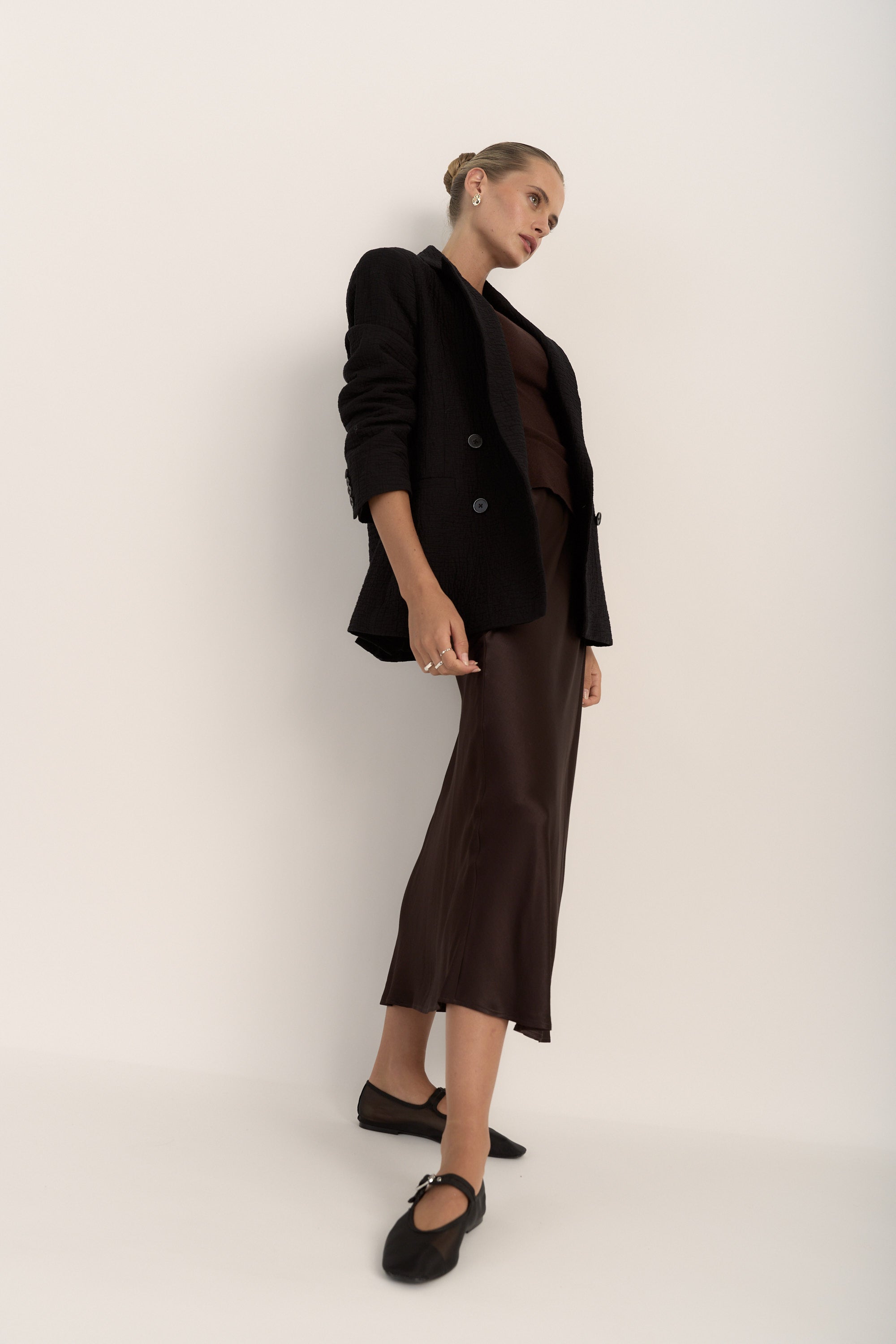 A woman poses against a white background wearing Max’s The Slip Skirt in brown satin, paired with a black blazer and black flats. Her hair is tied back, and one hand rests relaxed by her side.