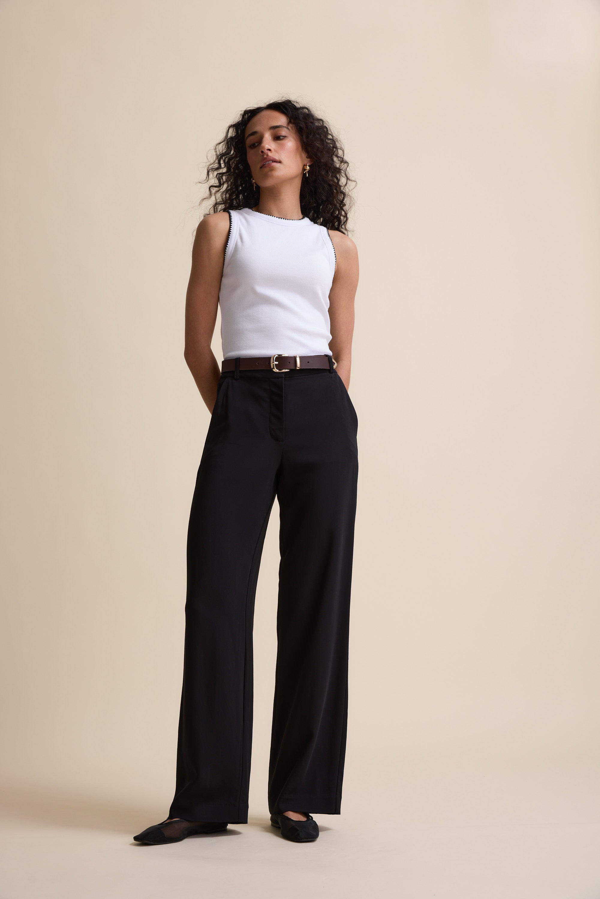A woman with curly hair stands against a beige background, hands in her pockets, wearing Max’s The Wide Leg Pant in a viscose blend, a white sleeveless top, black belt, and black shoes.