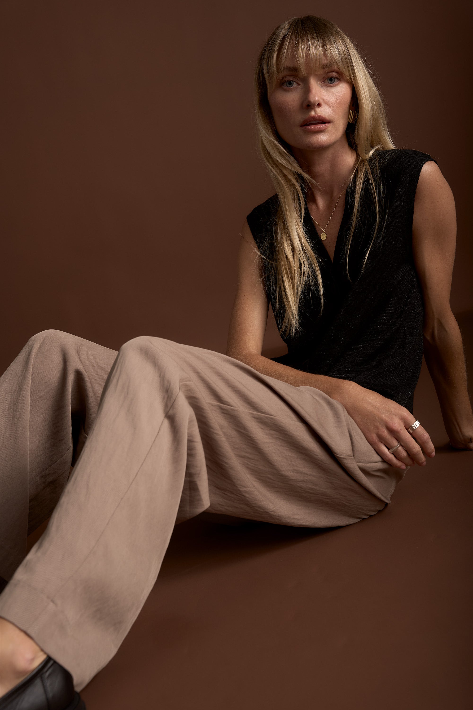 Woman in Max's The Ultimate Wide Leg Pants sits on a brown background in a black sleeveless top.