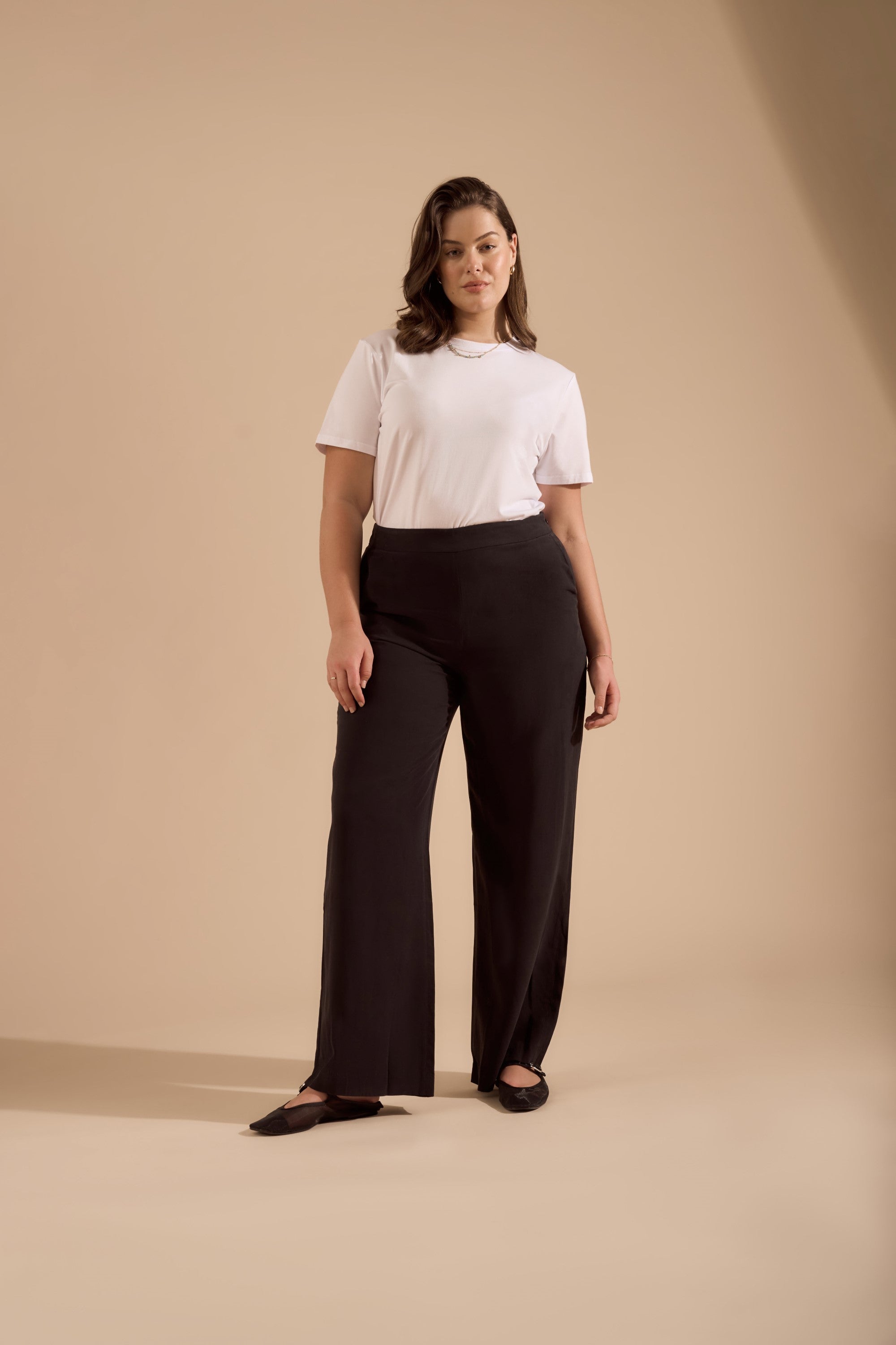 A woman with wavy brown hair and a neutral expression stands confidently in Max’s The Ultimate Wide Leg Pant and a white tee, paired with black flats, against a beige background. The high-waisted pants are made from sustainable fabrics.