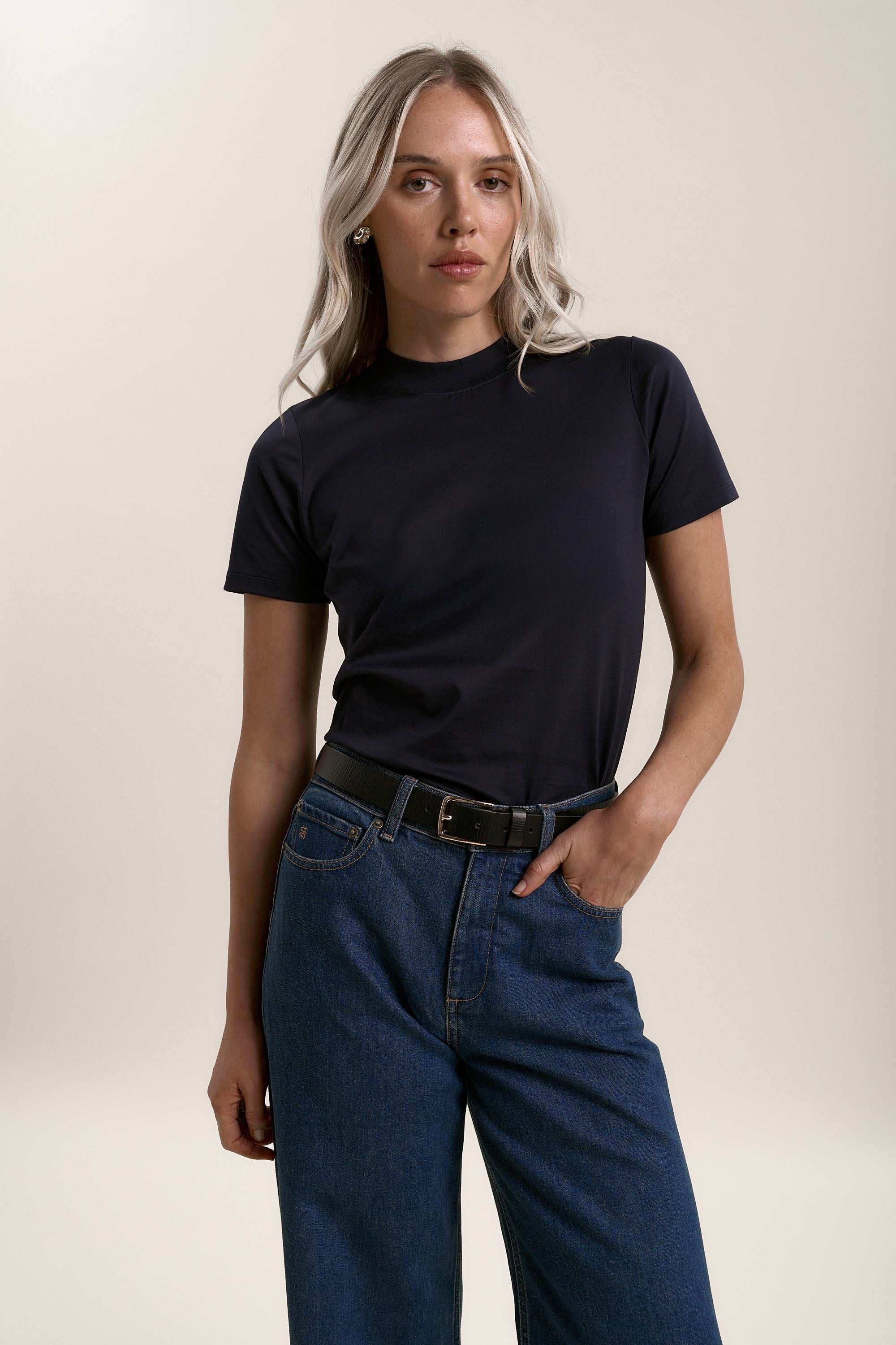 A person with long blonde hair wears the Max Erina High Neck Cotton Tee tucked into high-waisted blue jeans with a black belt, standing against a plain light background and looking at the camera with a neutral expression.