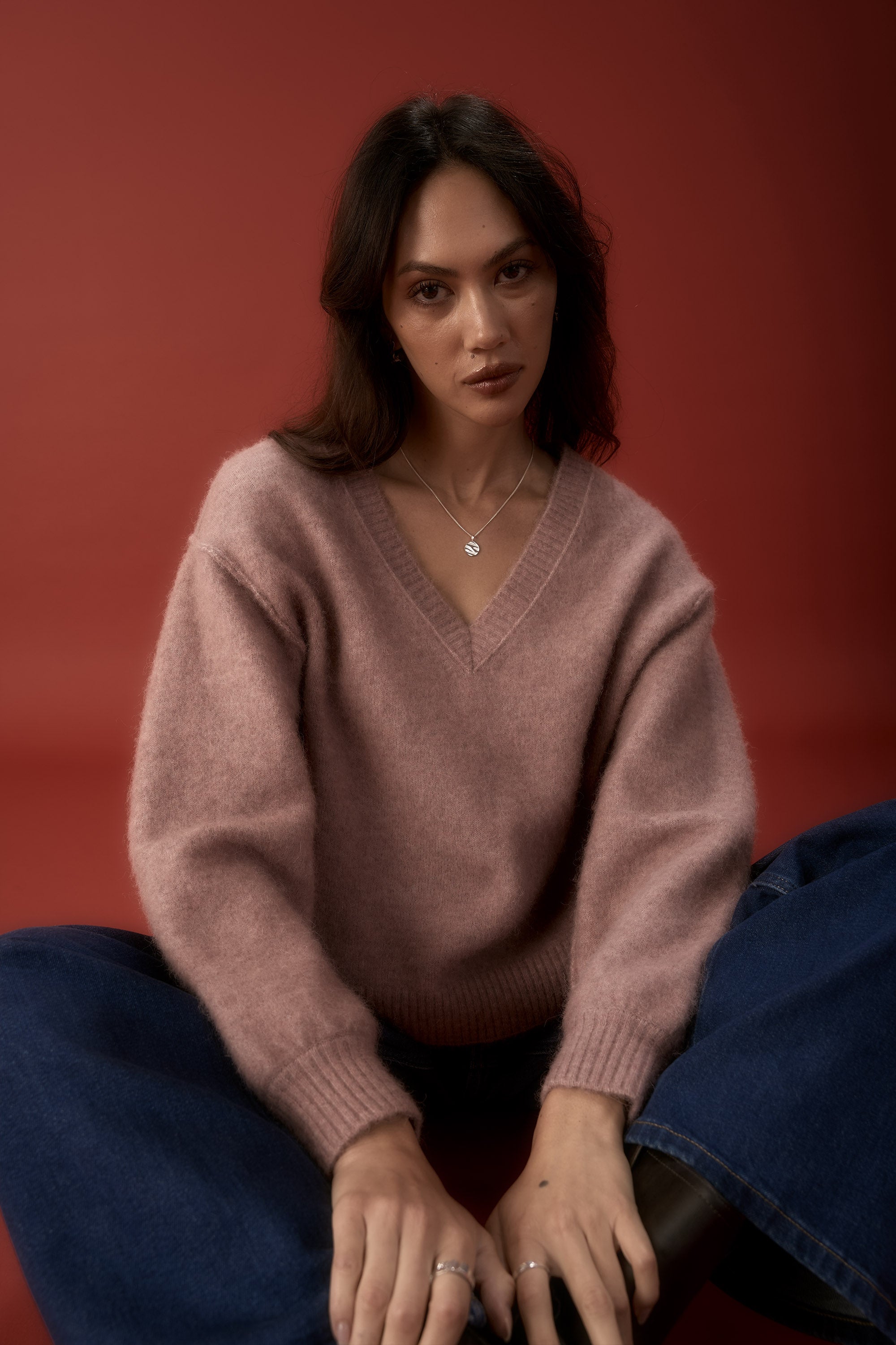 Woman in Max’s Luxe Whisper V Neck Sweater and blue jeans sits against a red background.