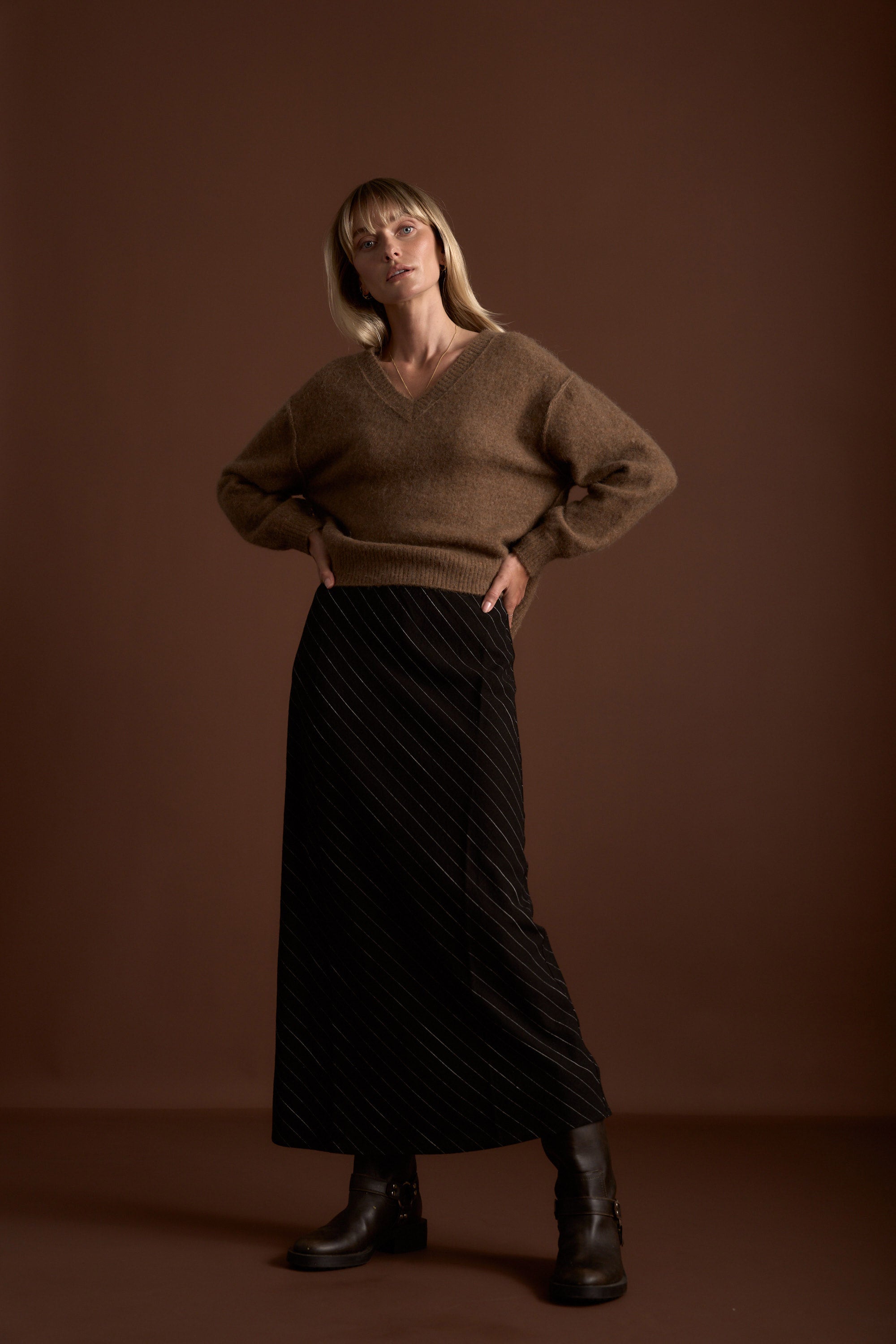 Woman in Max's Luxe Whisper V Neck Sweater, black skirt, and boots stands against brown.