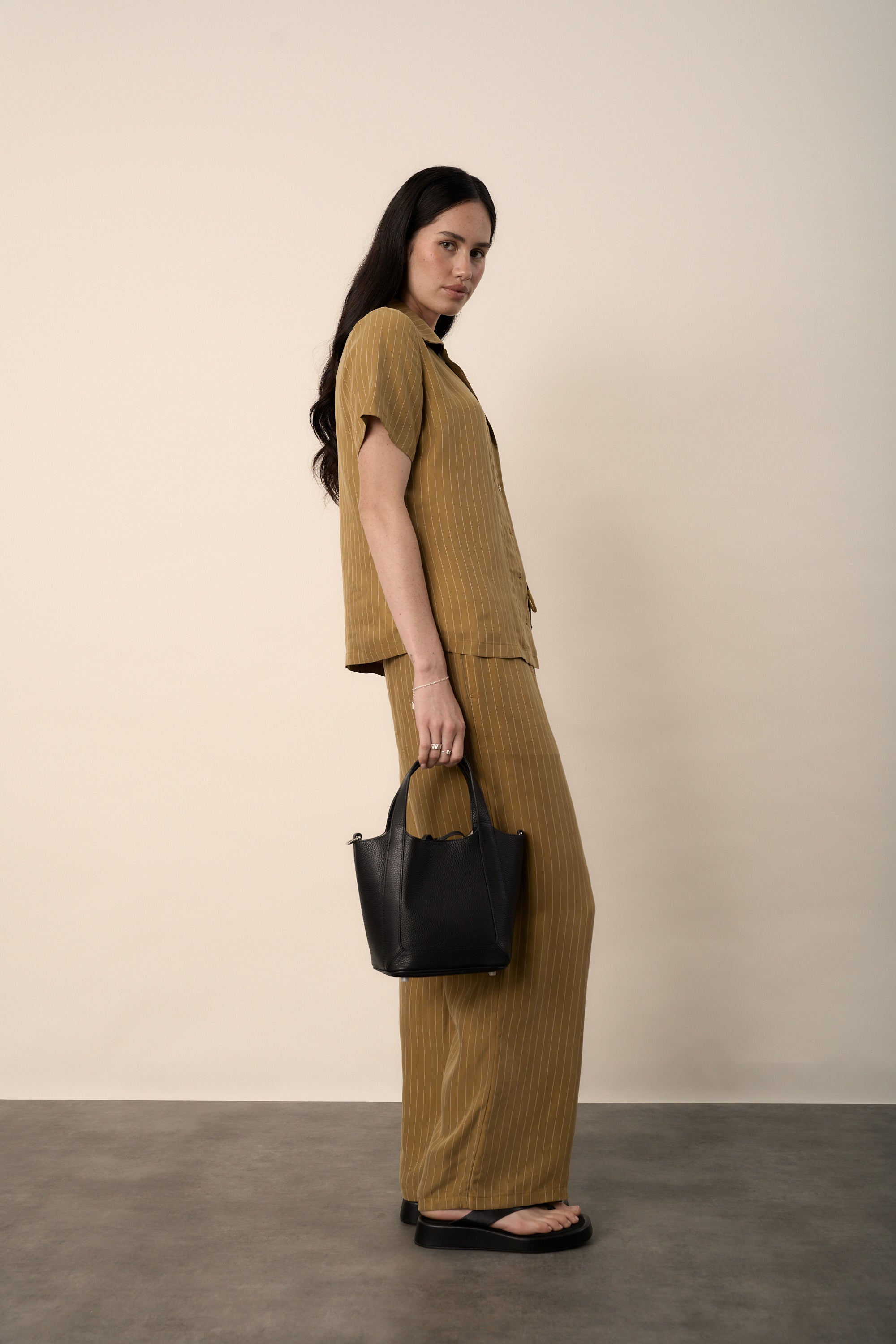 Woman in Amy Short Sleeve Shirt by Max holding black handbag, beige wall backdrop.