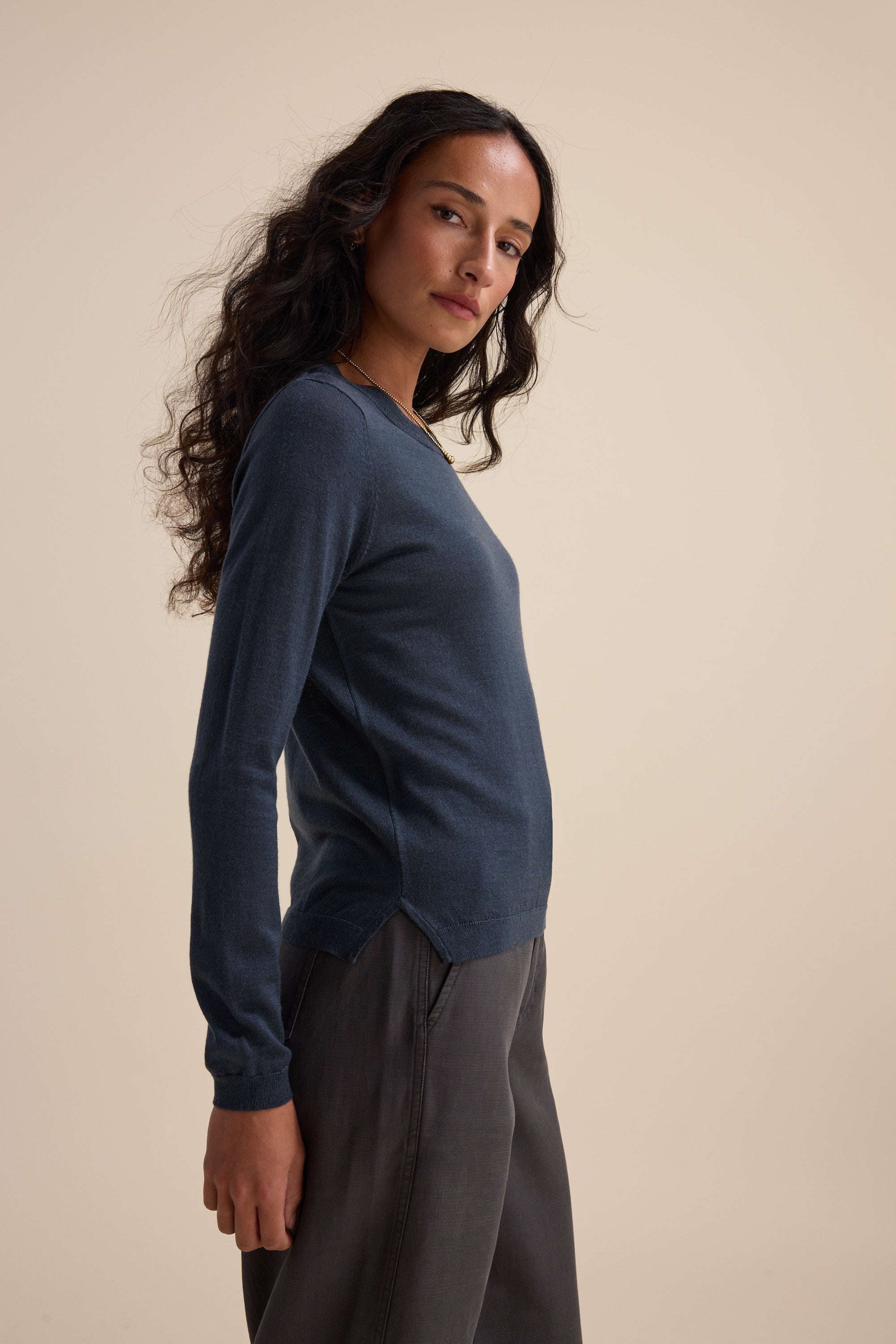 Woman in Max's Merino Silk Layering Top (blue) and gray pants, standing sideways on beige.