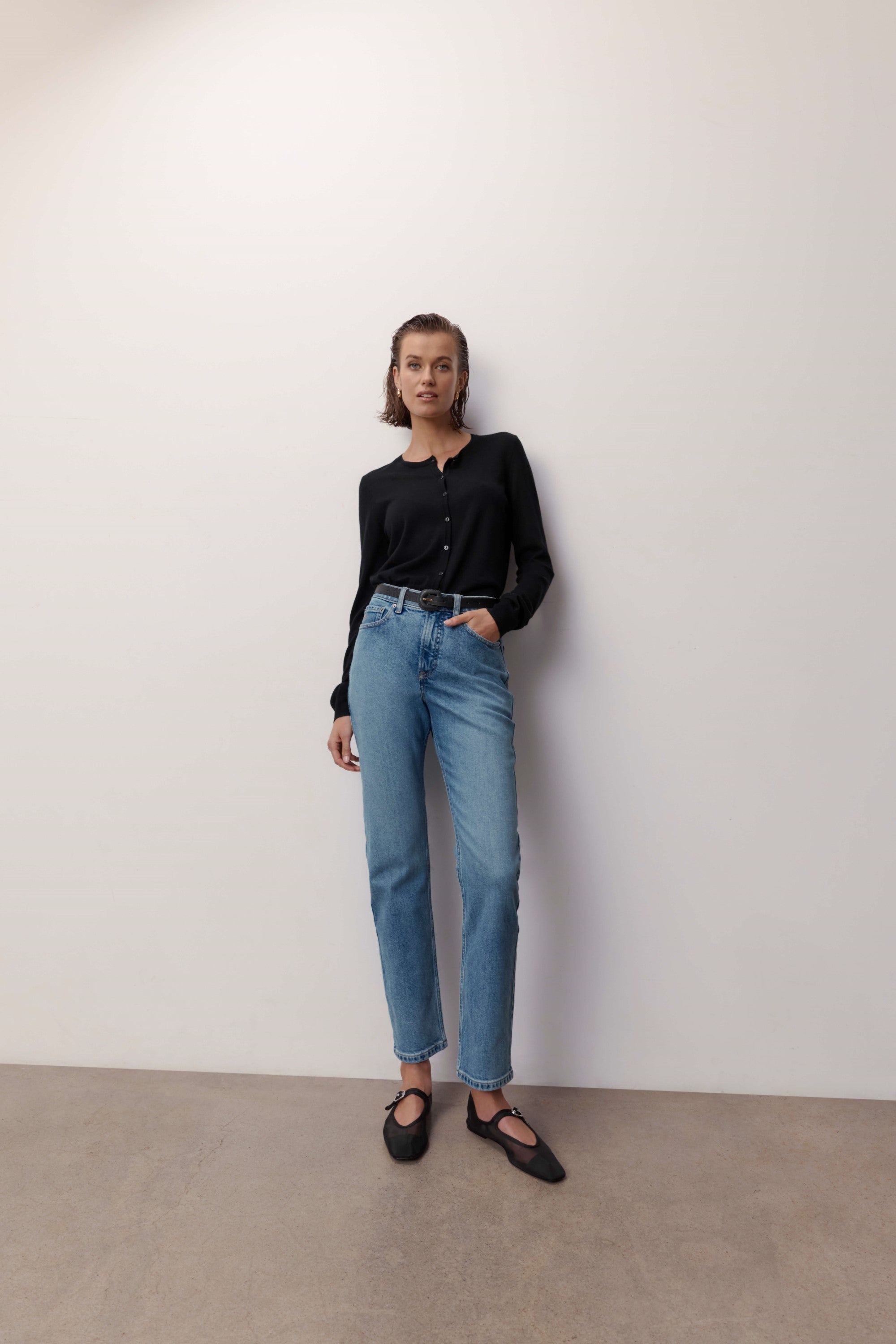 Woman in black blouse, Max Georgia Slim Jeans, and black shoes stands by a white wall.