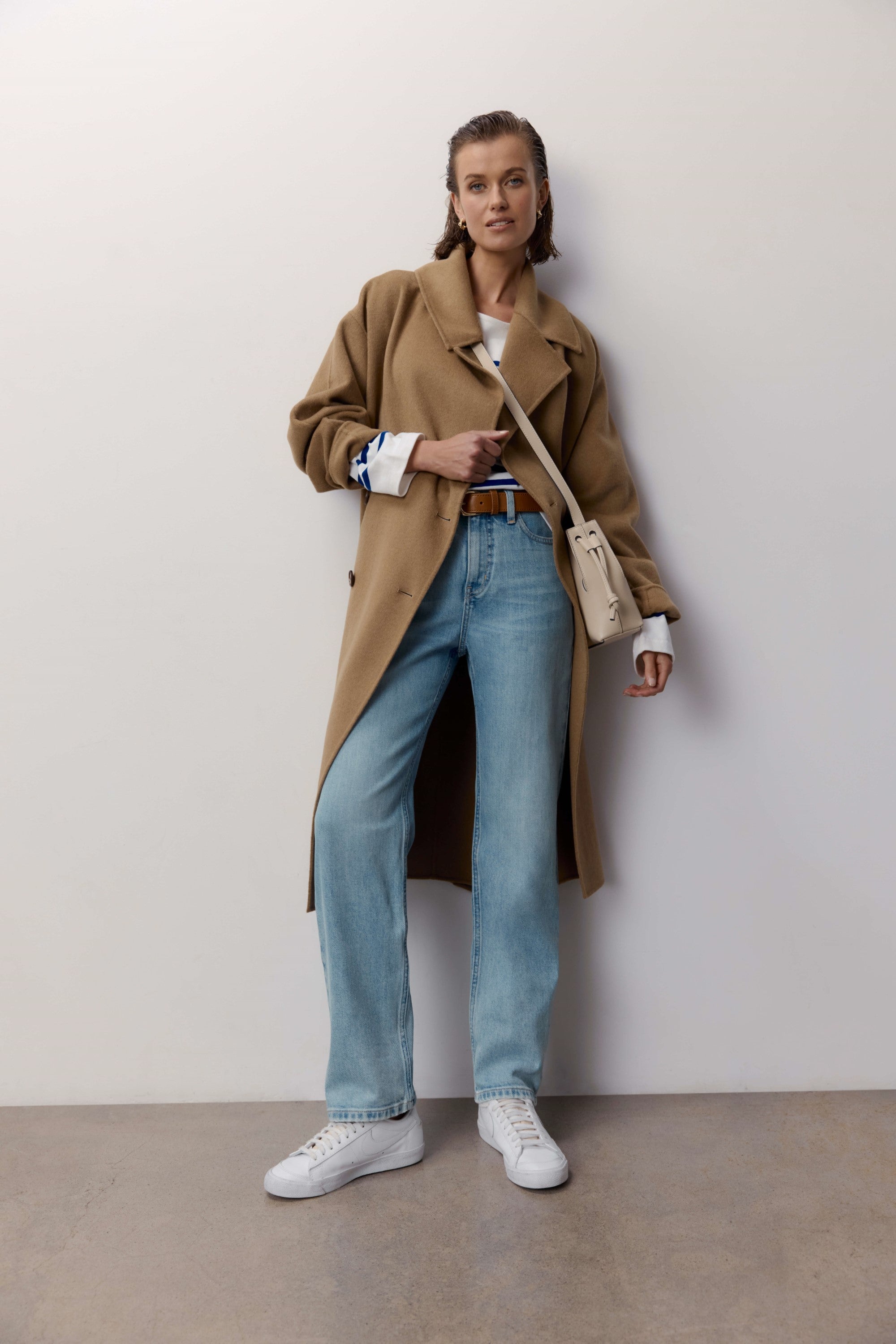 Woman in tan coat, white top, Max Arizona Straight Jeans, white sneakers, and beige bag by wall.