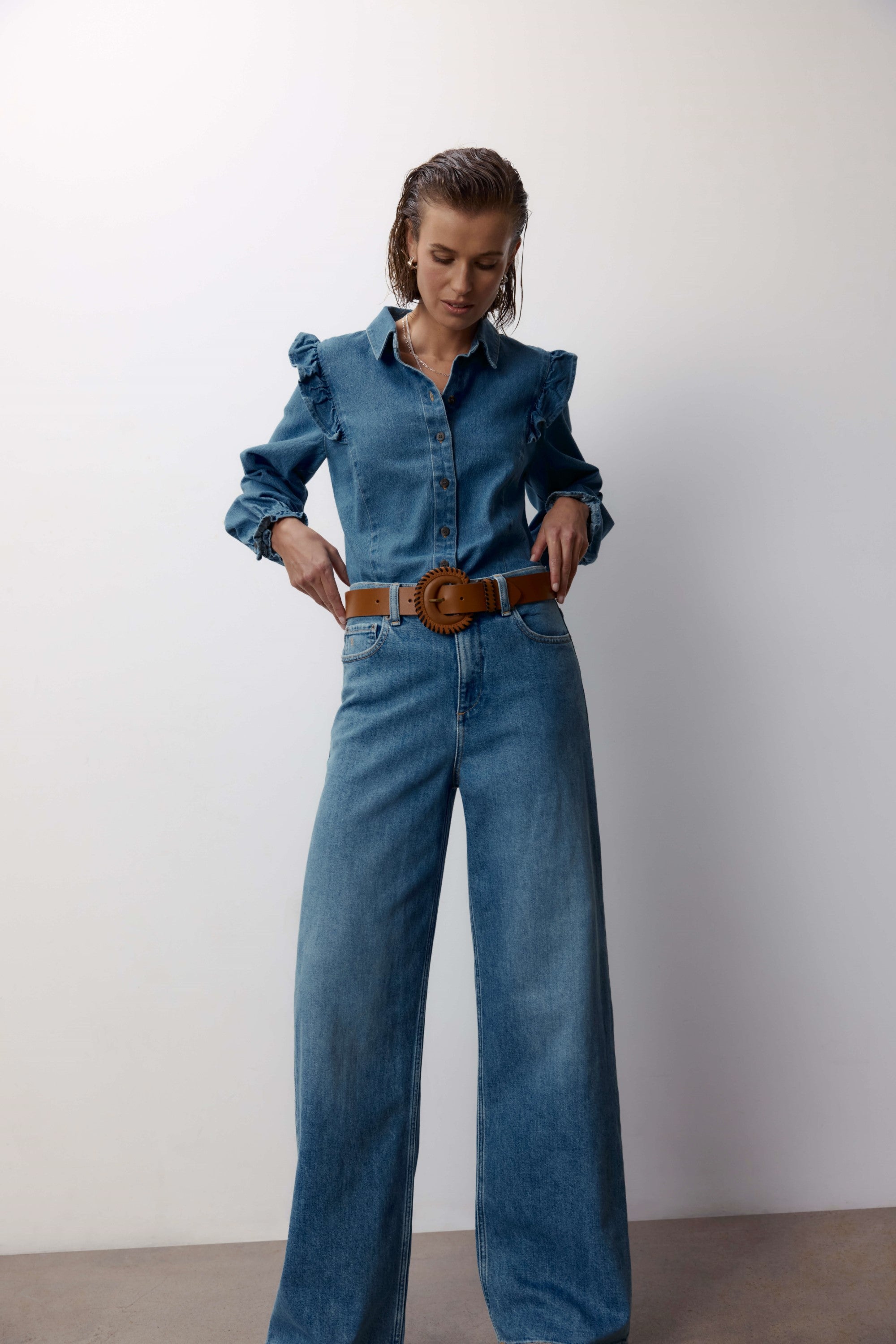 A woman in a denim shirt with ruffled shoulders and Max’s Nevada Wide Jeans stands against a plain background, looking down as she adjusts a tan belt at her waist.