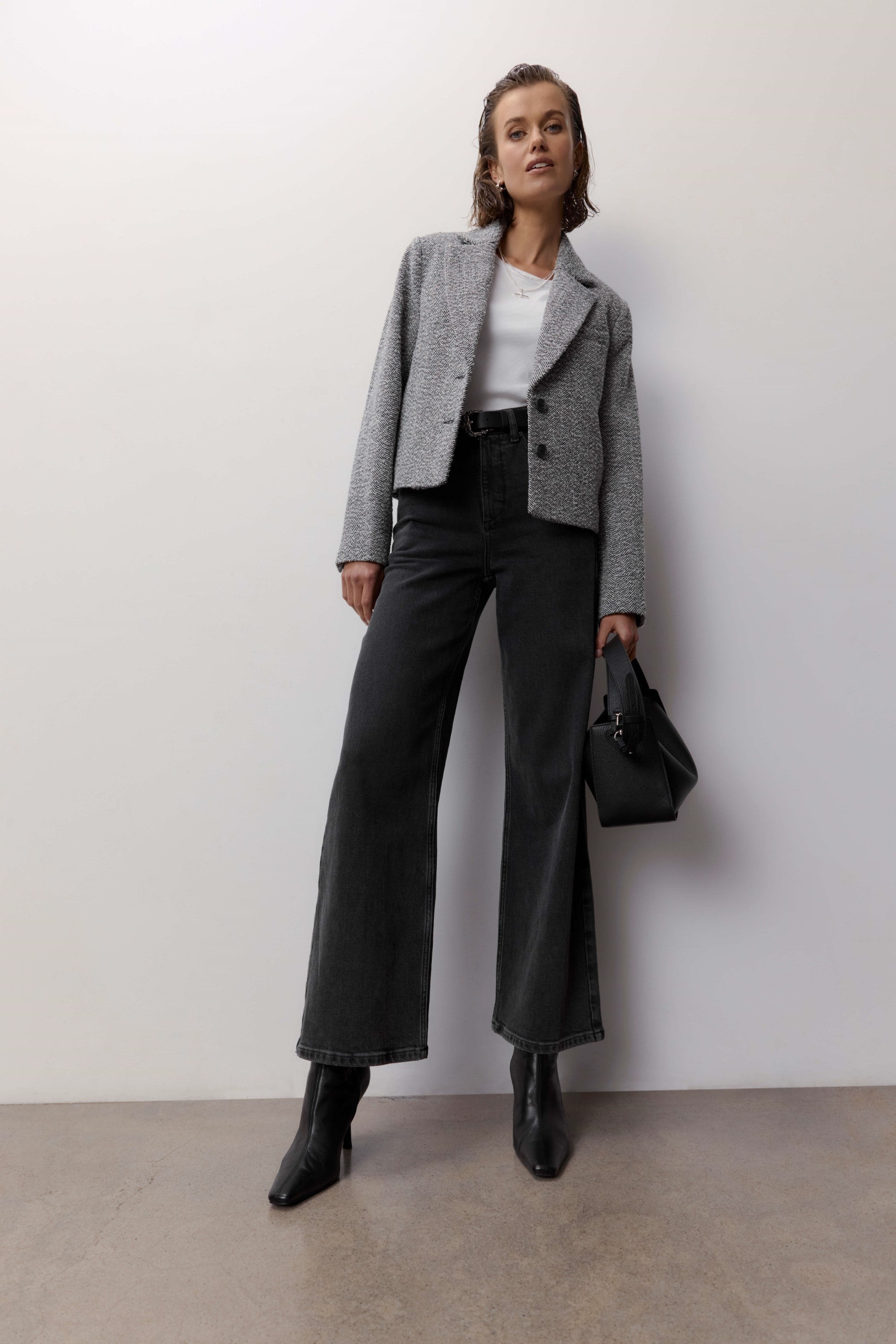 A woman stands against a plain wall wearing Max’s Nevada Wide Jeans in black sustainable denim, paired with a grey blazer, white top, black handbag, and ankle boots as she looks confidently at the camera.