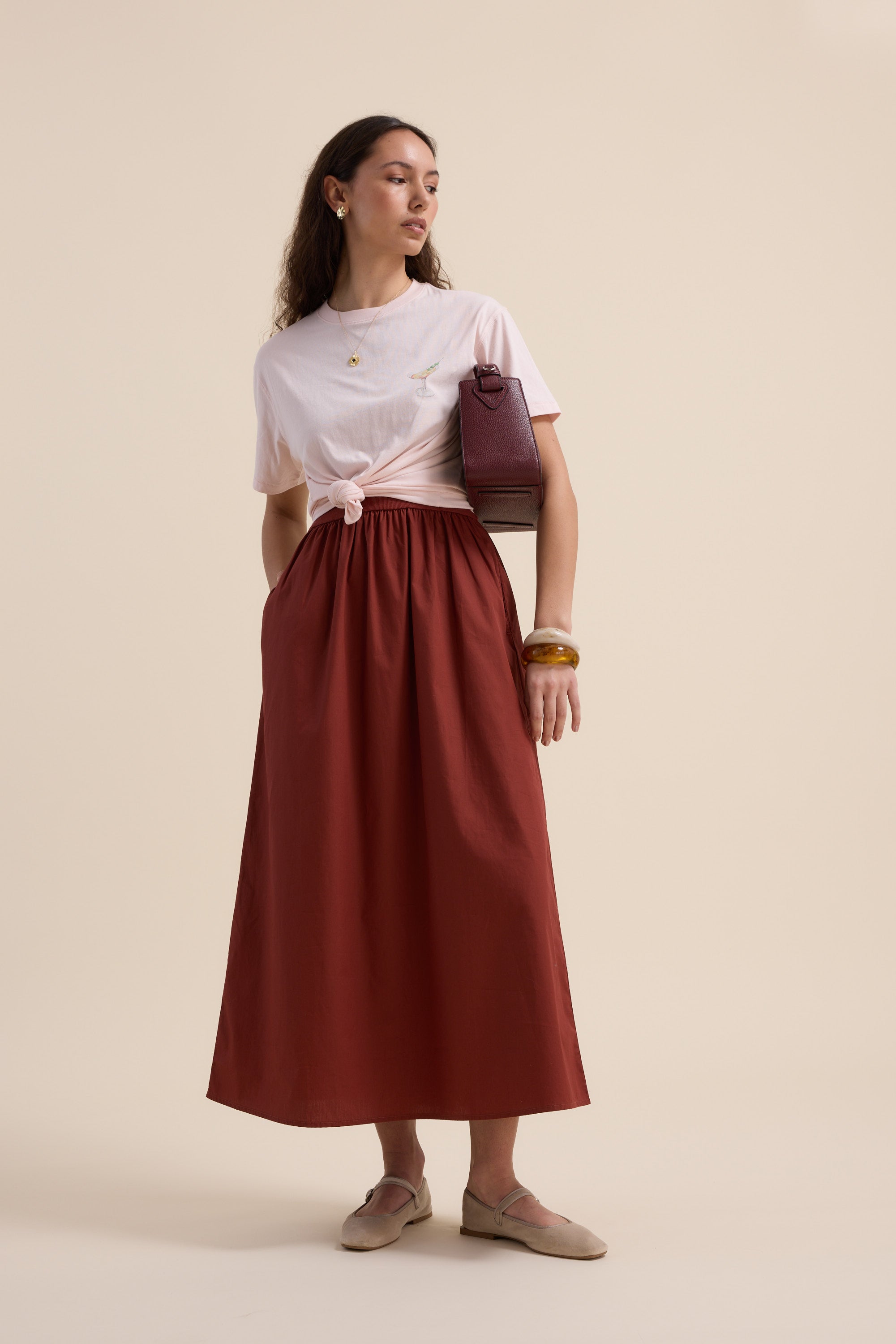 A woman with long brown hair and gold jewelry poses against a beige backdrop in Max's The One And Done Dress, styled simply with beige flats and a maroon handbag.