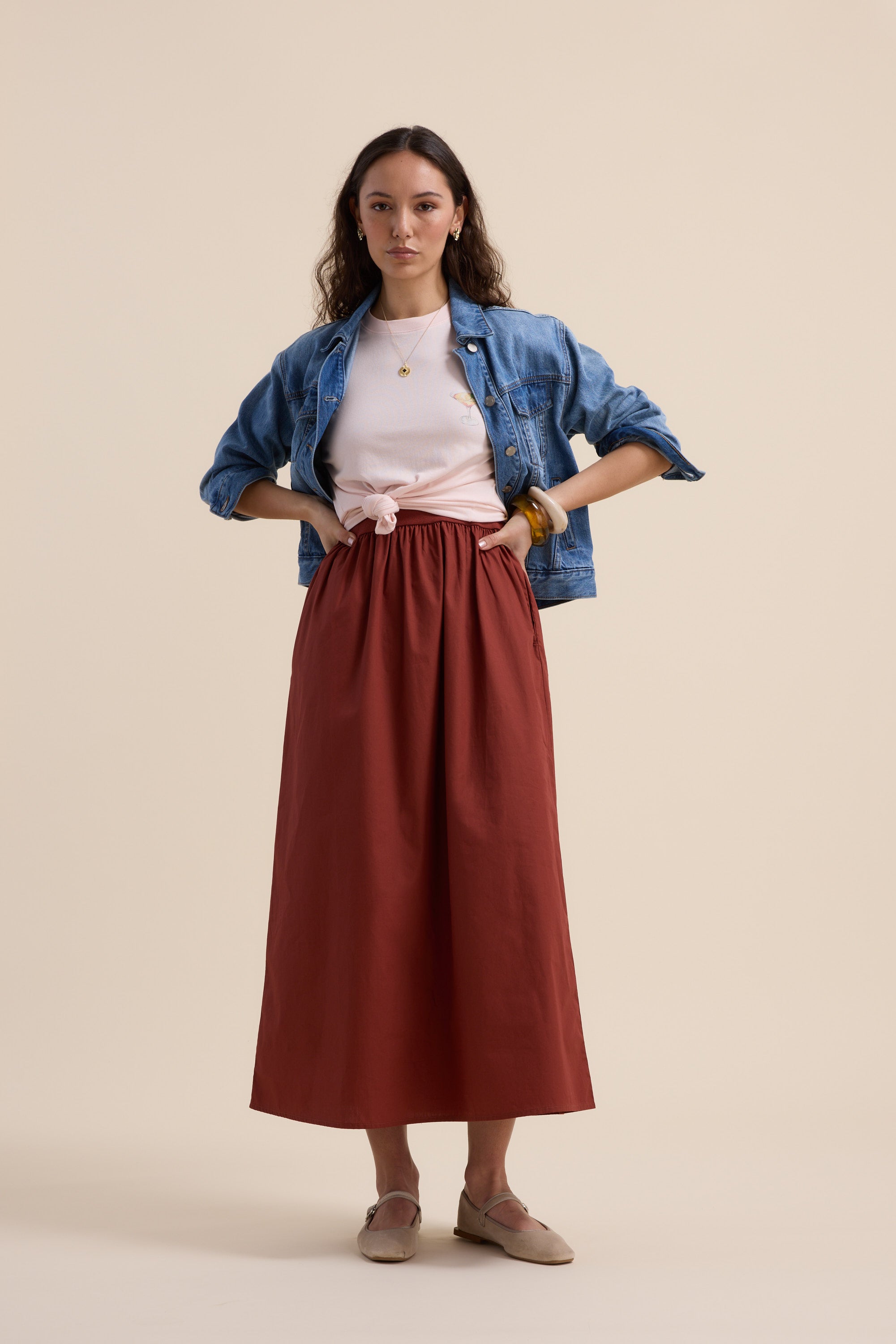 A woman poses against a beige background in Max’s The One And Done Dress—a long rust-colored organic cotton midi—styled with a white top, denim jacket, beige flats, and a gold necklace for an effortlessly versatile look.