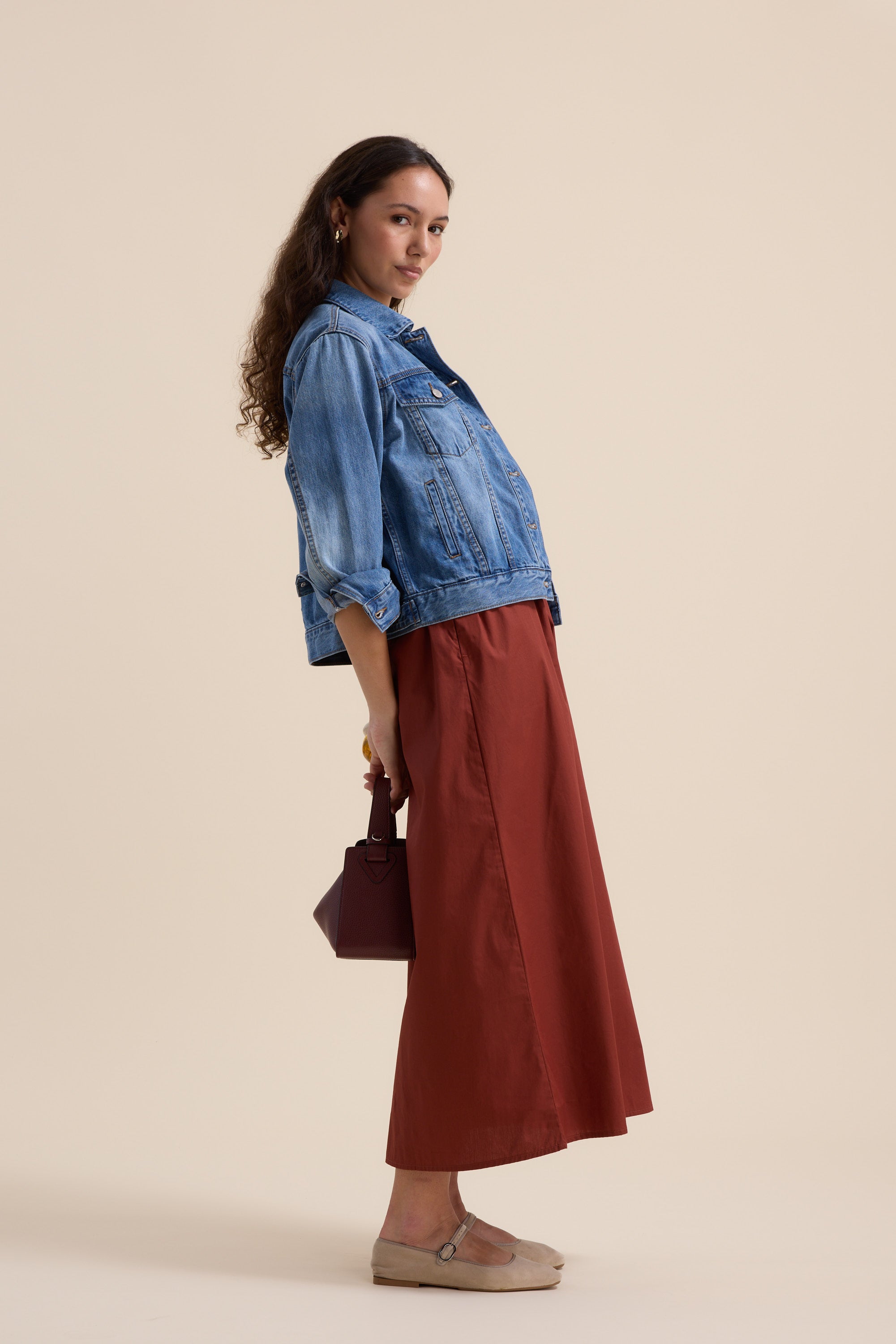 A woman with long curly hair stands in profile against a beige background, wearing Max’s The One And Done Dress in rust, styled with beige shoes and a small dark handbag, striking a relaxed pose.