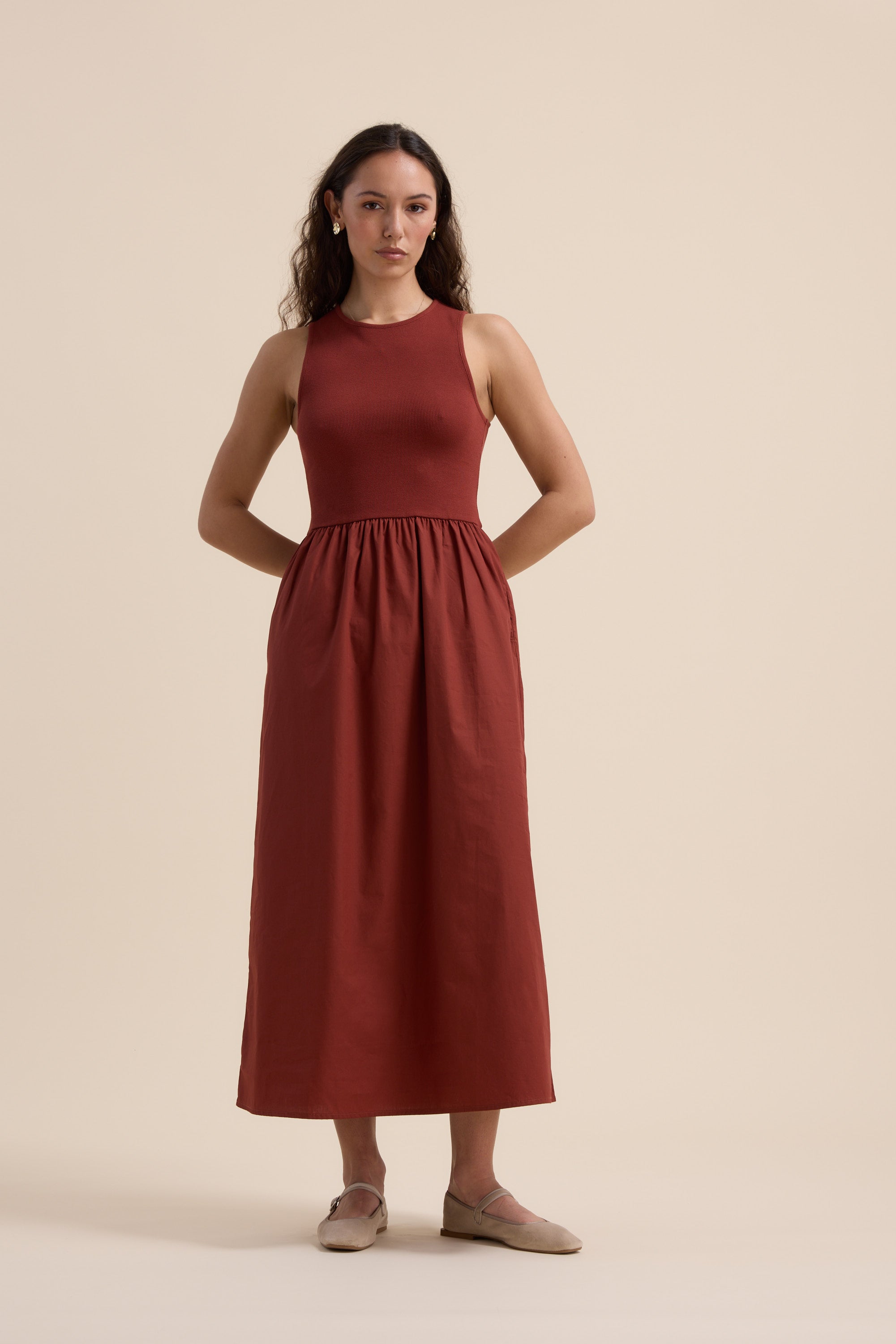 A woman with long, wavy hair stands against a beige backdrop, wearing Max’s The One And Done Dress—a rust-toned organic cotton midi with a fitted bodice and gathered skirt—styled simply with neutral flats, hands behind her back.