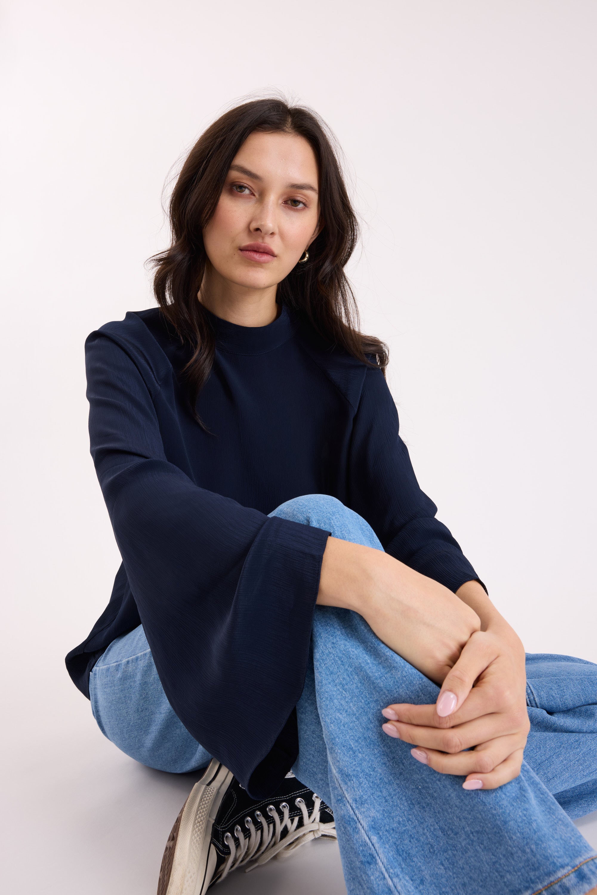 Woman wearing Max Lindsay Textured Blouse and blue jeans, sitting cross-legged, facing camera.