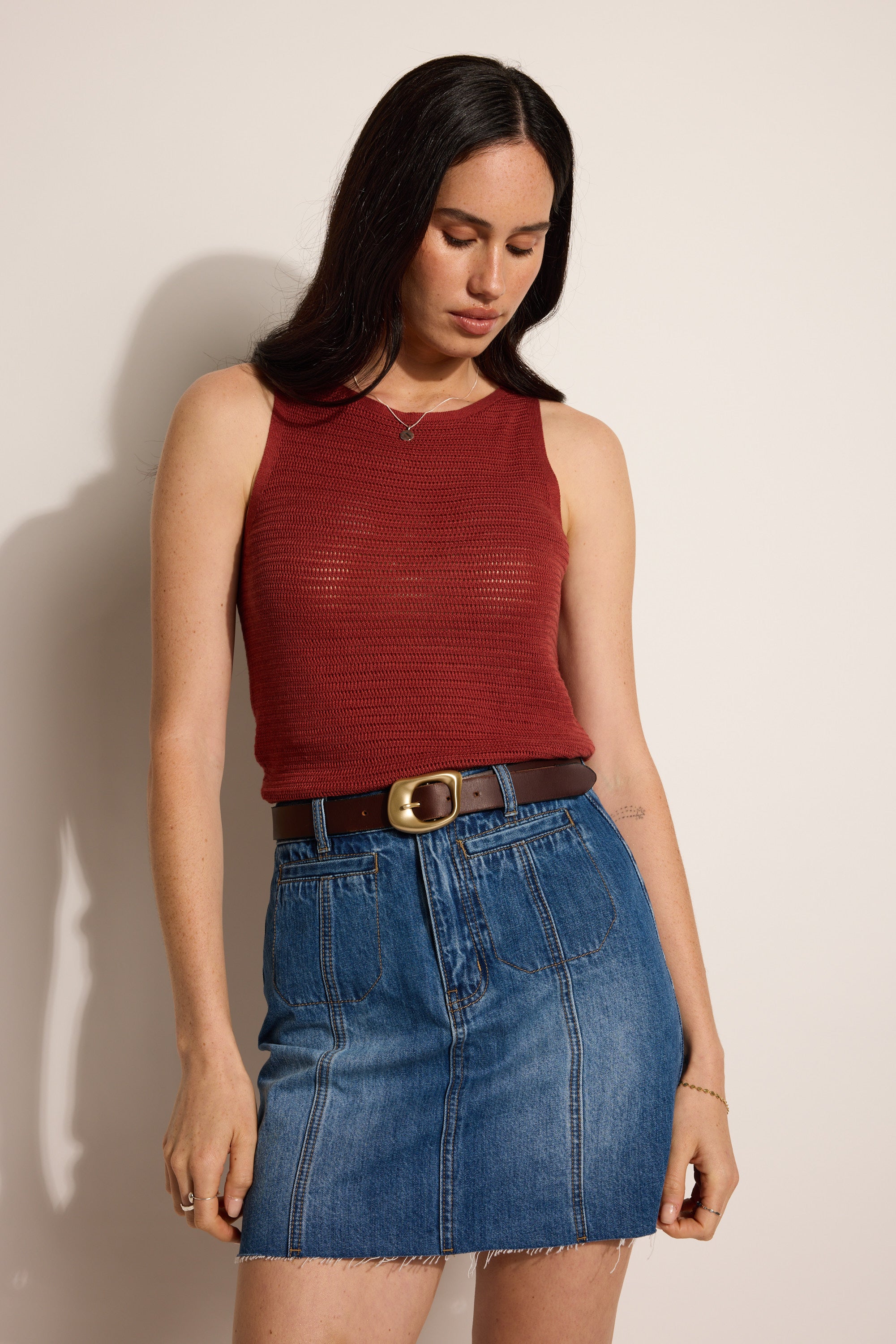 A woman with long dark hair wears the Elementary Cove Knit Tank in red, paired with a brown belt and blue denim skirt. She stands against a light background, looking down with her arms relaxed at her sides.