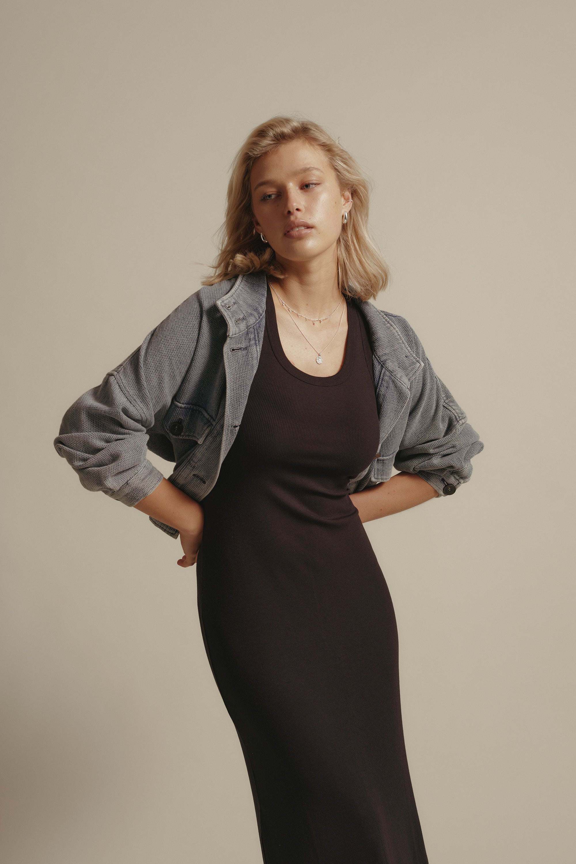 A blonde woman wears the Elementary Basics Tank Slip by Elementary, a black midi dress, paired with a cropped gray denim jacket. She stands against a light background, hands on hips, with simple jewelry and a calm expression.