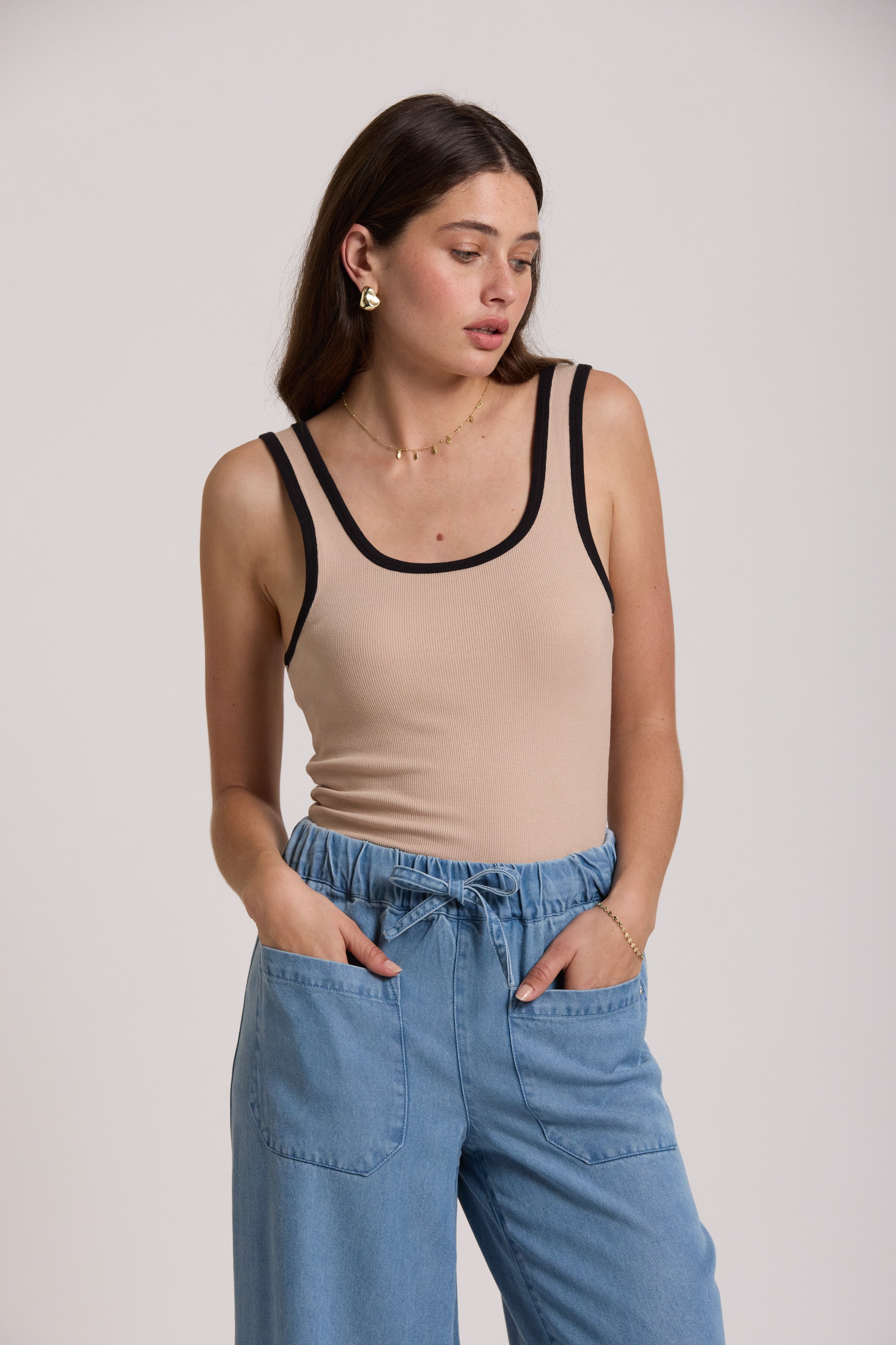 Woman wears Max's Contrast Trim Tank in beige with blue jeans, hands in pockets, looking aside.