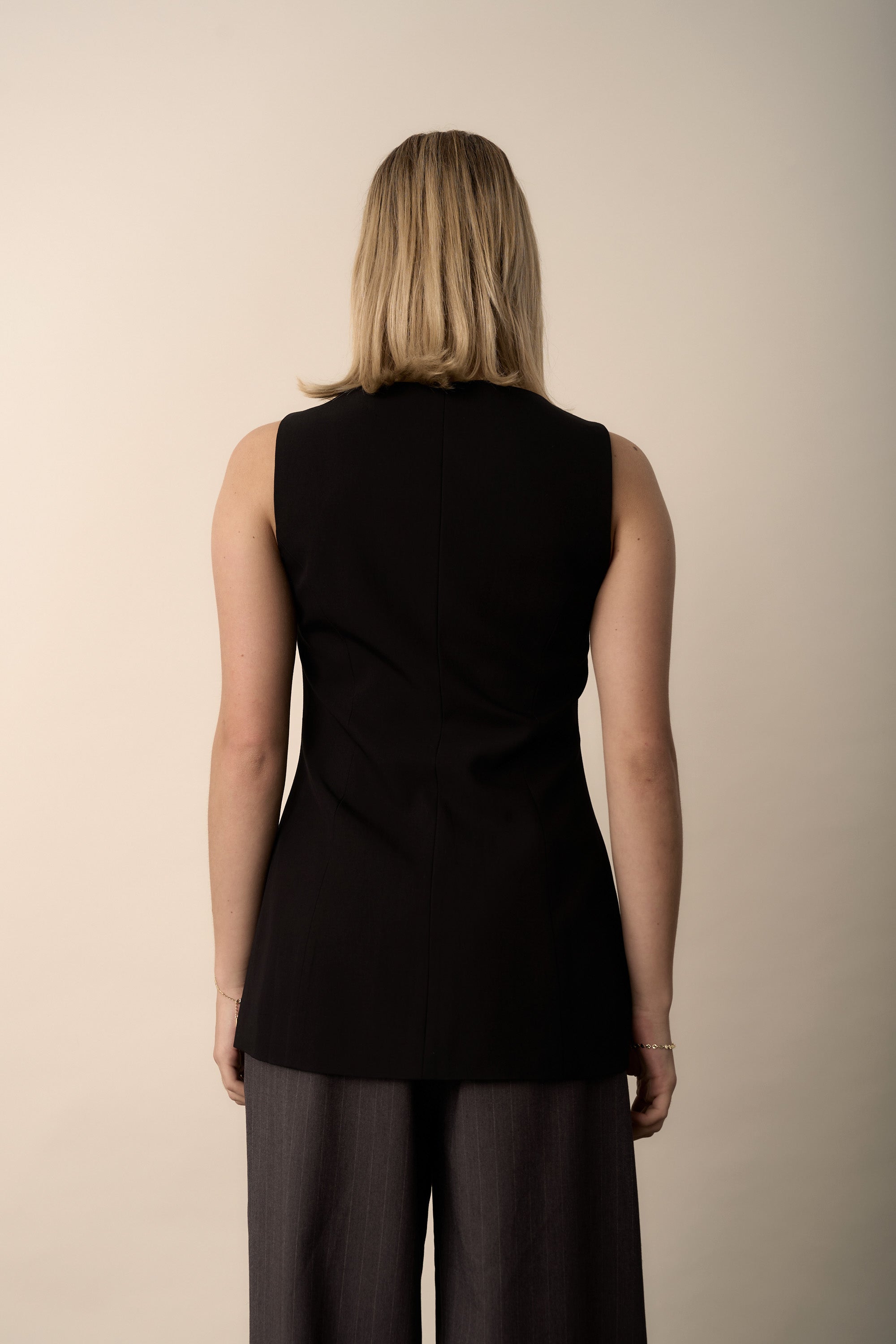 A woman with blonde hair faces away, wearing the Max Button Front Vest and gray pants.