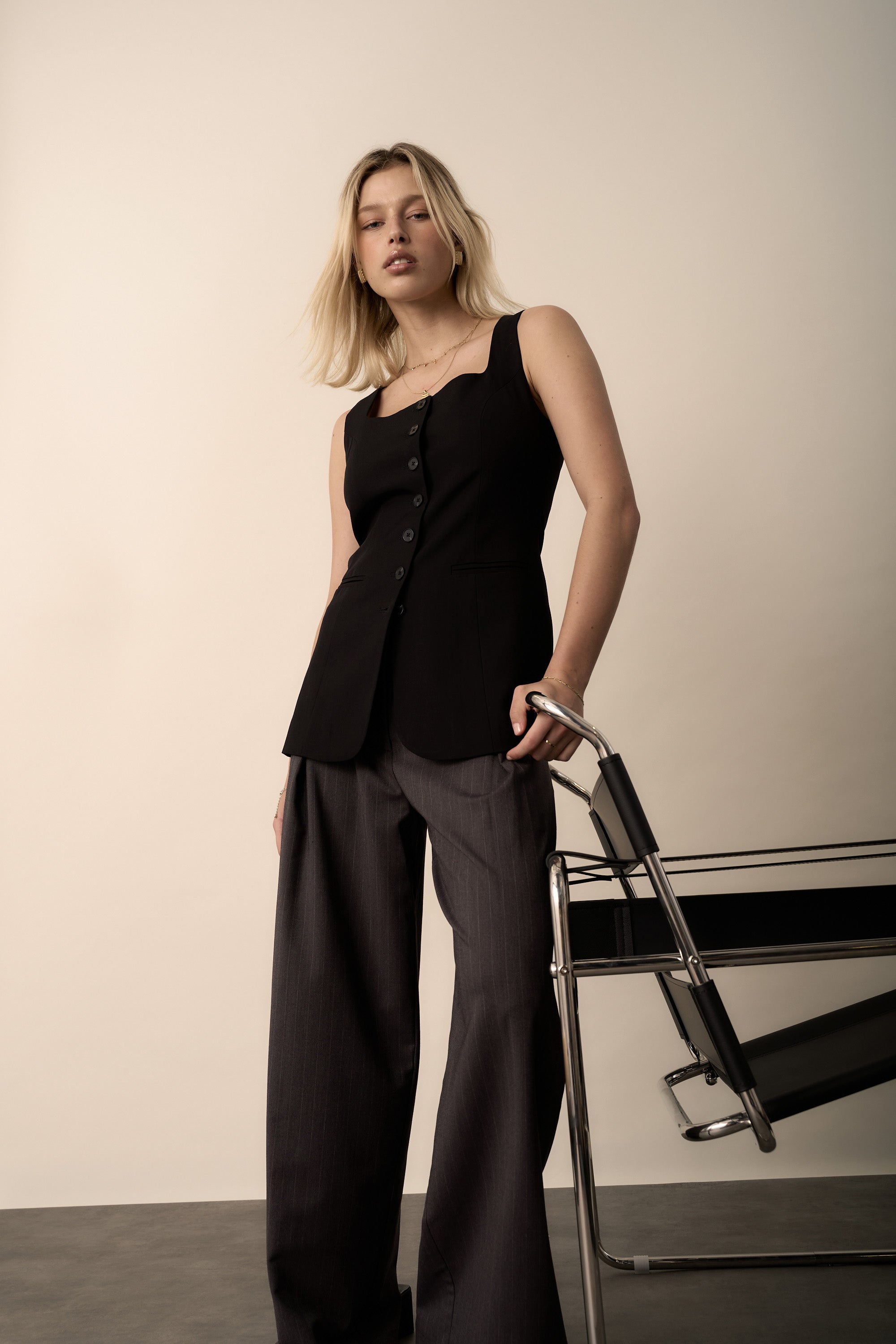 Woman wears Max’s The Button Front Vest, standing by a modern chair in a studio.