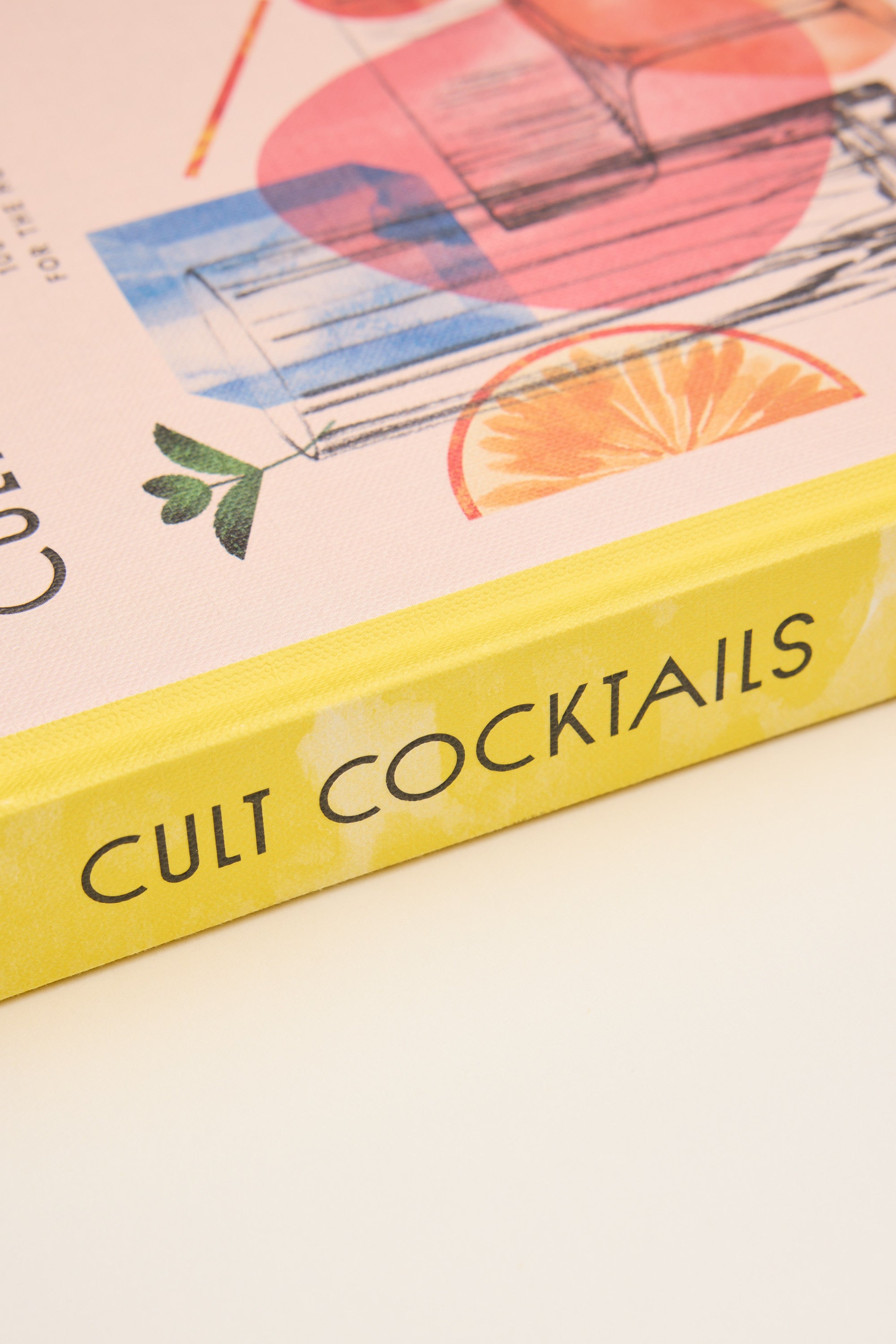 Close-up of the Cult Cocktails Book by Other, with a yellow spine and pastel cover decorated with cocktail infographics, fruit slices, and leaves—an inspiring resource for home bartenders—resting on a white surface.