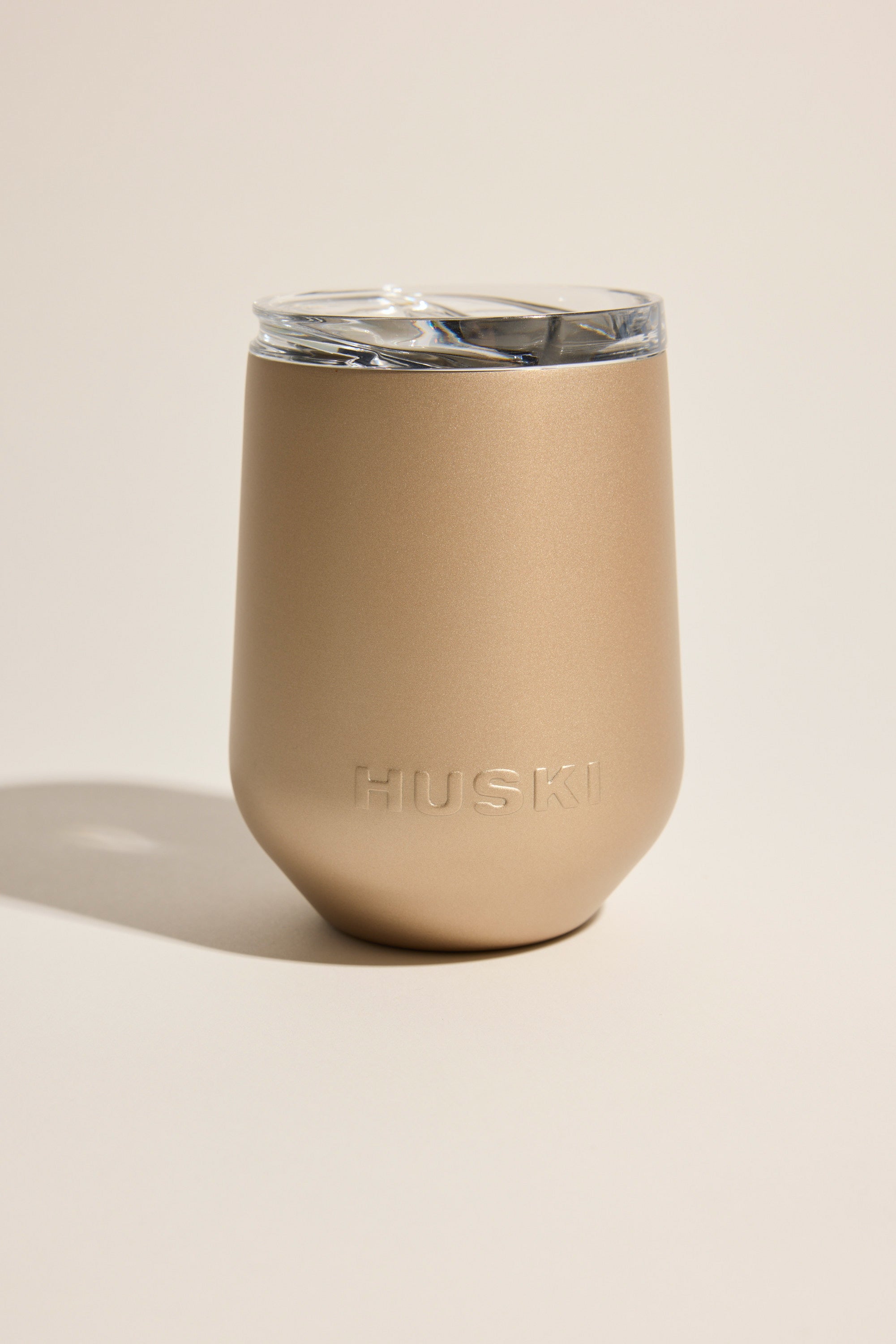 The Huski Wine Tumbler 2.0 by Huski is a gold, matte-finish insulated cup with a clear lid and embossed branding on the front, shown against a neutral background.