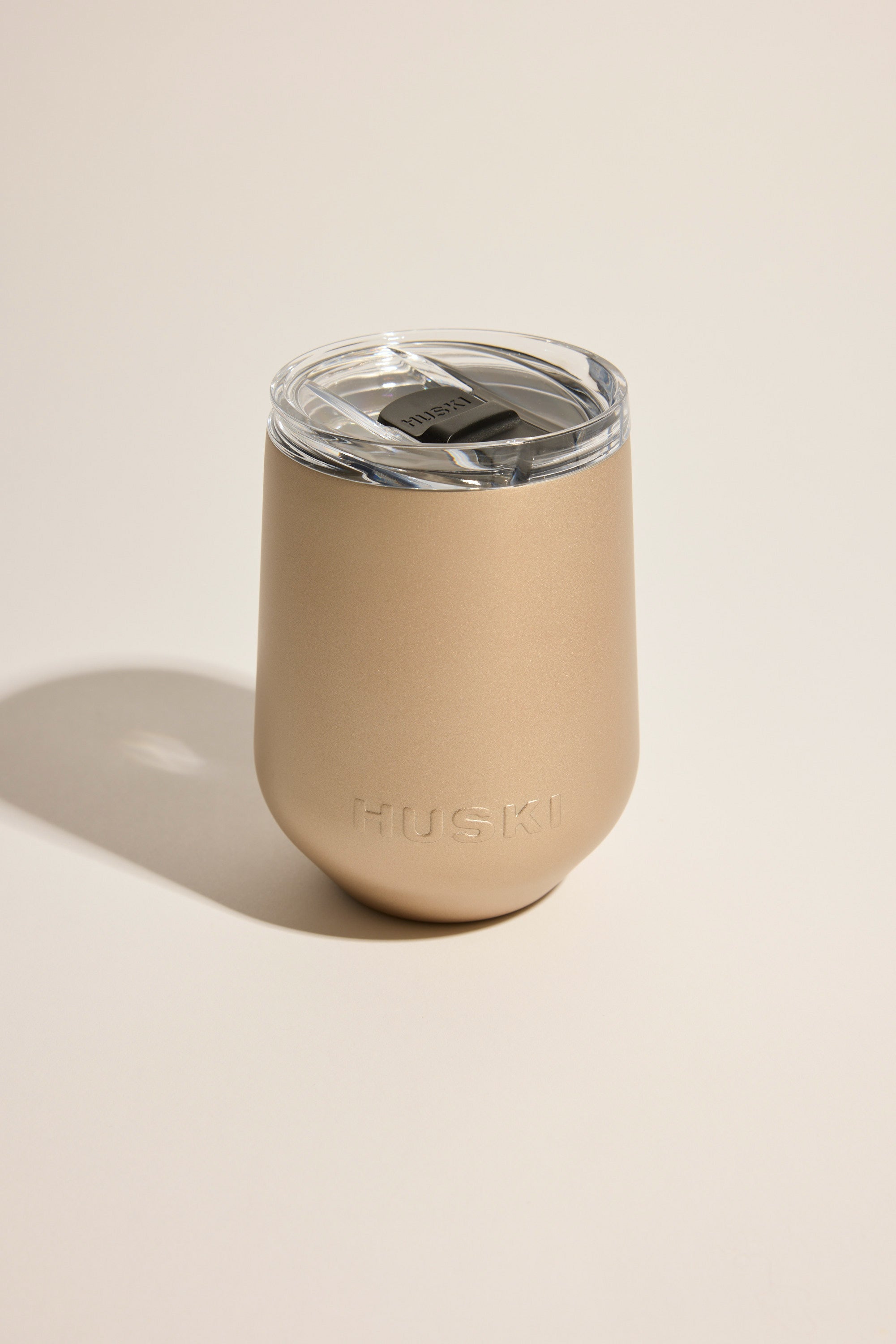 A gold stainless steel Huski Wine Tumbler 2.0 with a clear plastic lid, branded Huski, is placed on a light, neutral background.