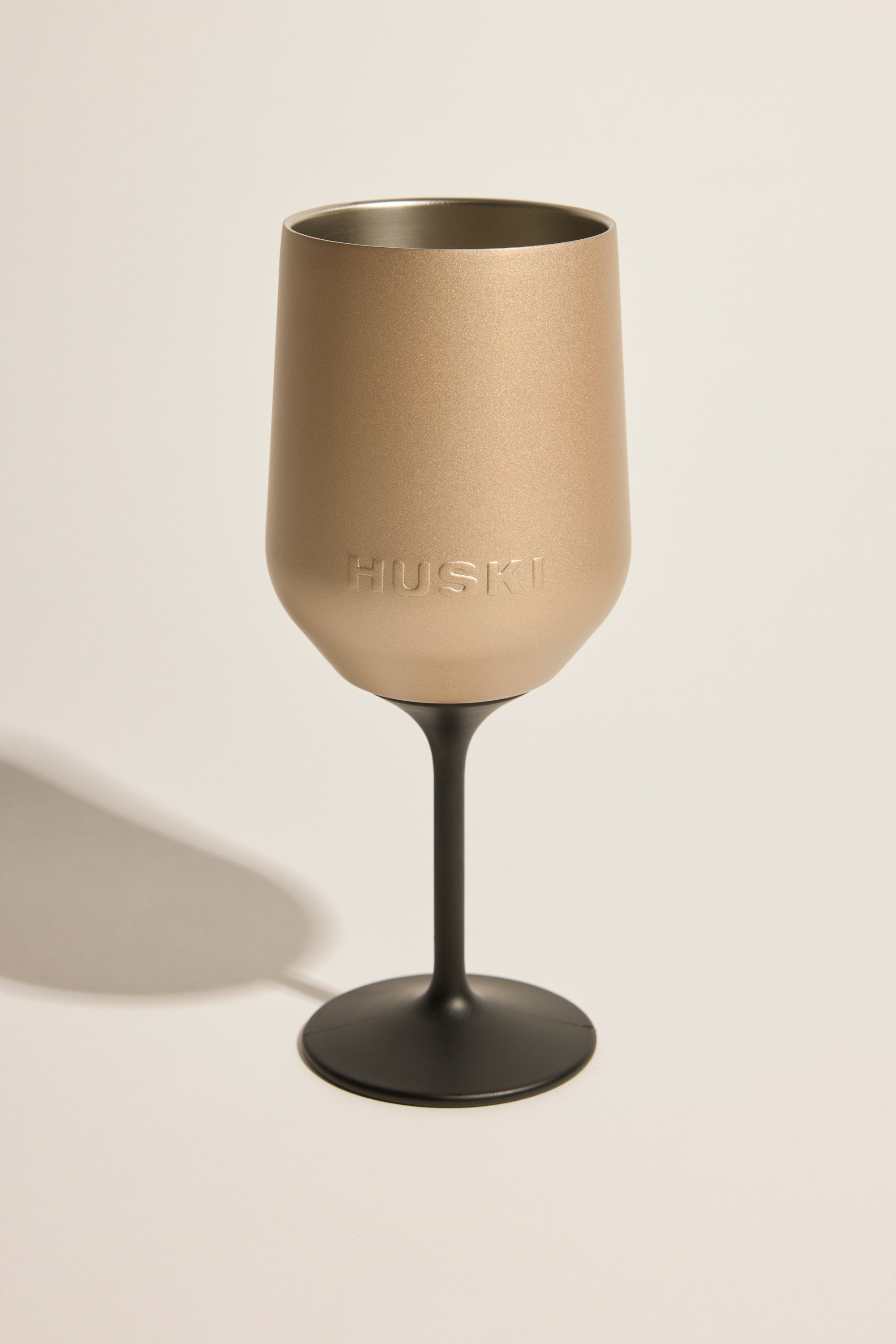 The Huski Wine Tumbler 2.0 by Huski is a metallic gold insulated tumbler with "HUSKI" embossed on the side, featuring a black stem and base, set against a plain light background.