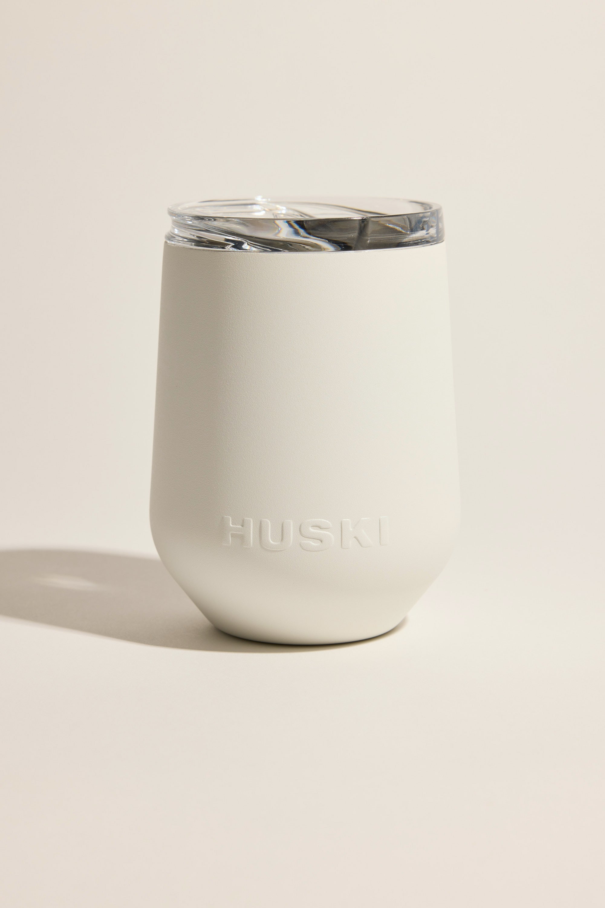 The Huski Wine Tumbler 2.0 by Huski, in white matte-finish stainless steel with a clear plastic lid and raised HUSKI logo on the front, sits against a plain, light background.