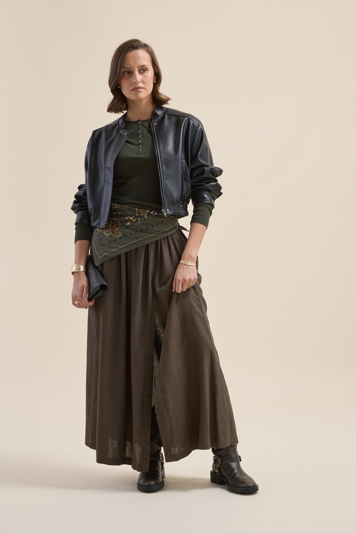 A woman wears a black jacket, Elementary Ribbed Henley in green, olive skirt, boots, and bag.