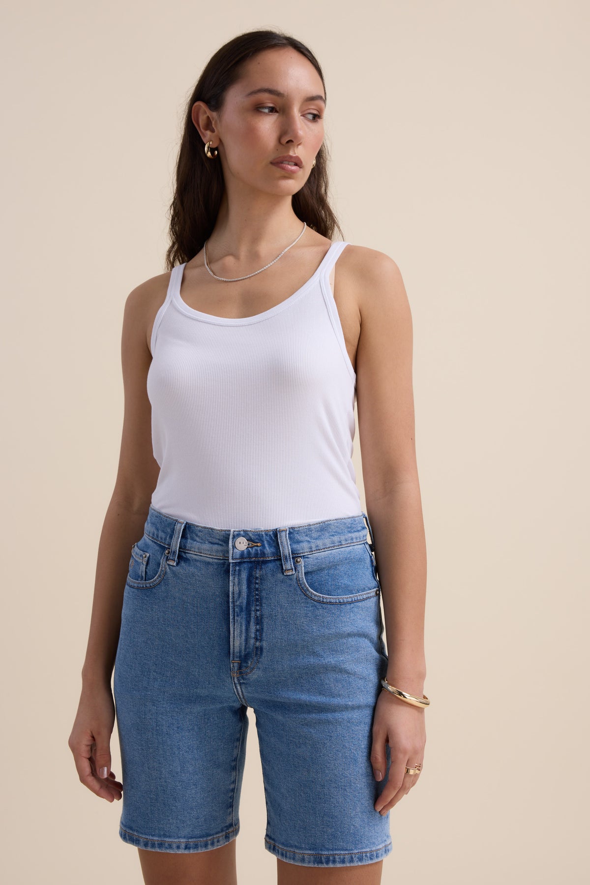 Woman wears Max's The Classic Denim Short with a white tank top against a beige background.