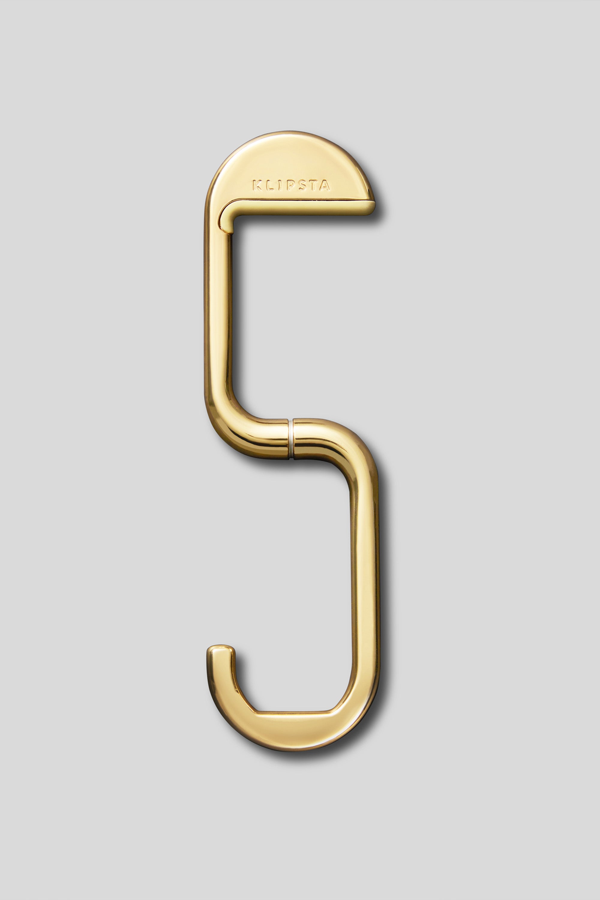 The Klipsta Flip Bag Hook by Klipsta is a gold metal handbag hook shaped like the number 5, engraved with "KLIPSTA" on the top, shown against a plain light gray background.