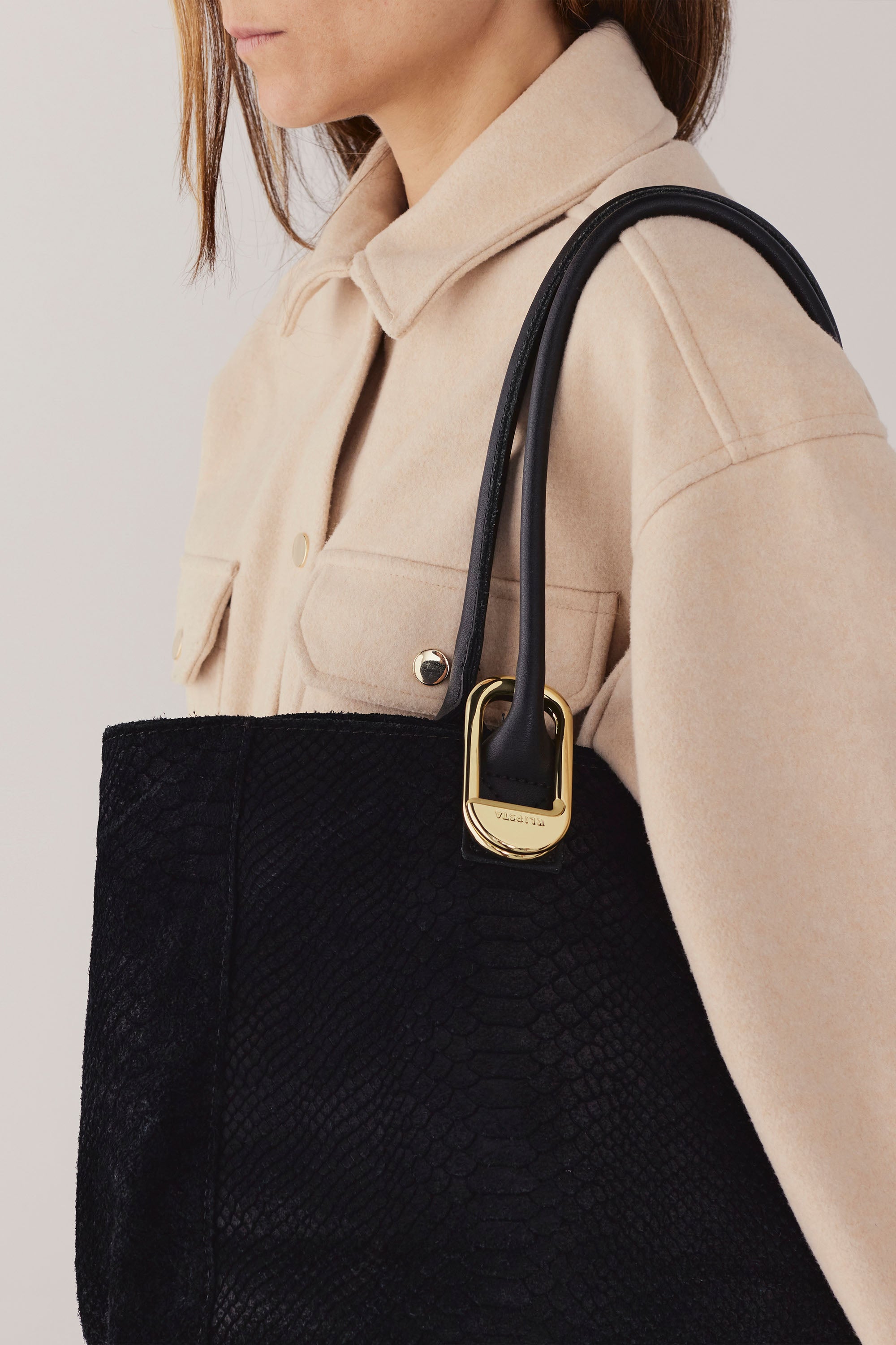 A woman wearing a beige button-up shirt carries a large black tote with gold hardware on her shoulder, featuring the Klipsta Flip Bag Hook by Klipsta as a stylish portable bag hanger. Only her upper body is visible.