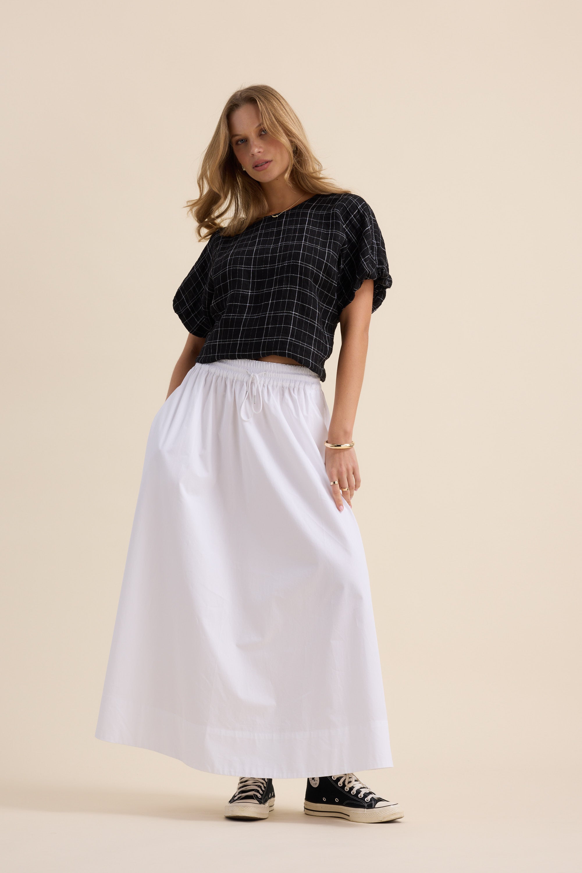 Woman in Max's The Bubble Hem Top, white skirt, and black sneakers poses against beige background.
