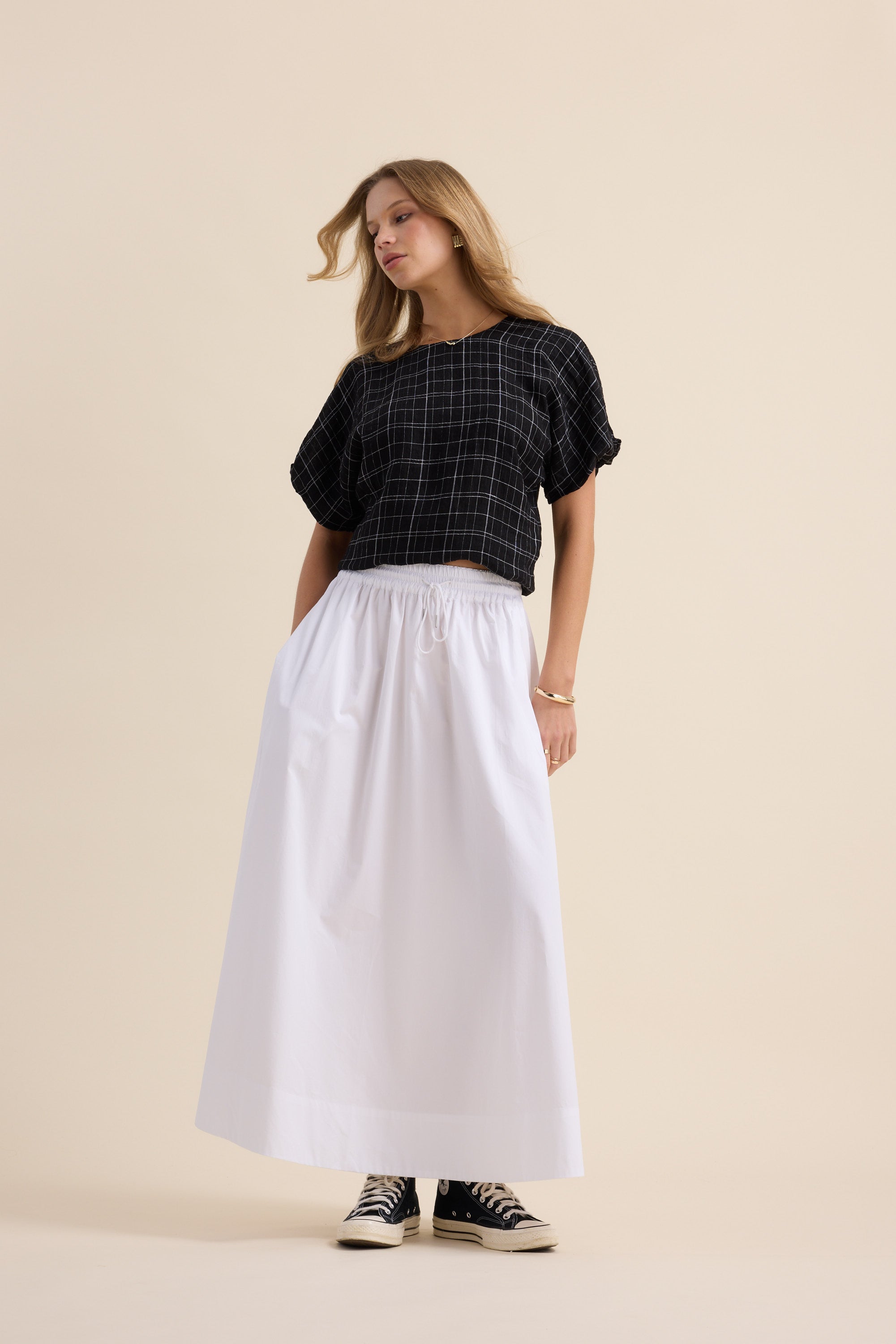 Woman in Max’s The Bubble Hem Top, white maxi skirt, and black sneakers against beige background.