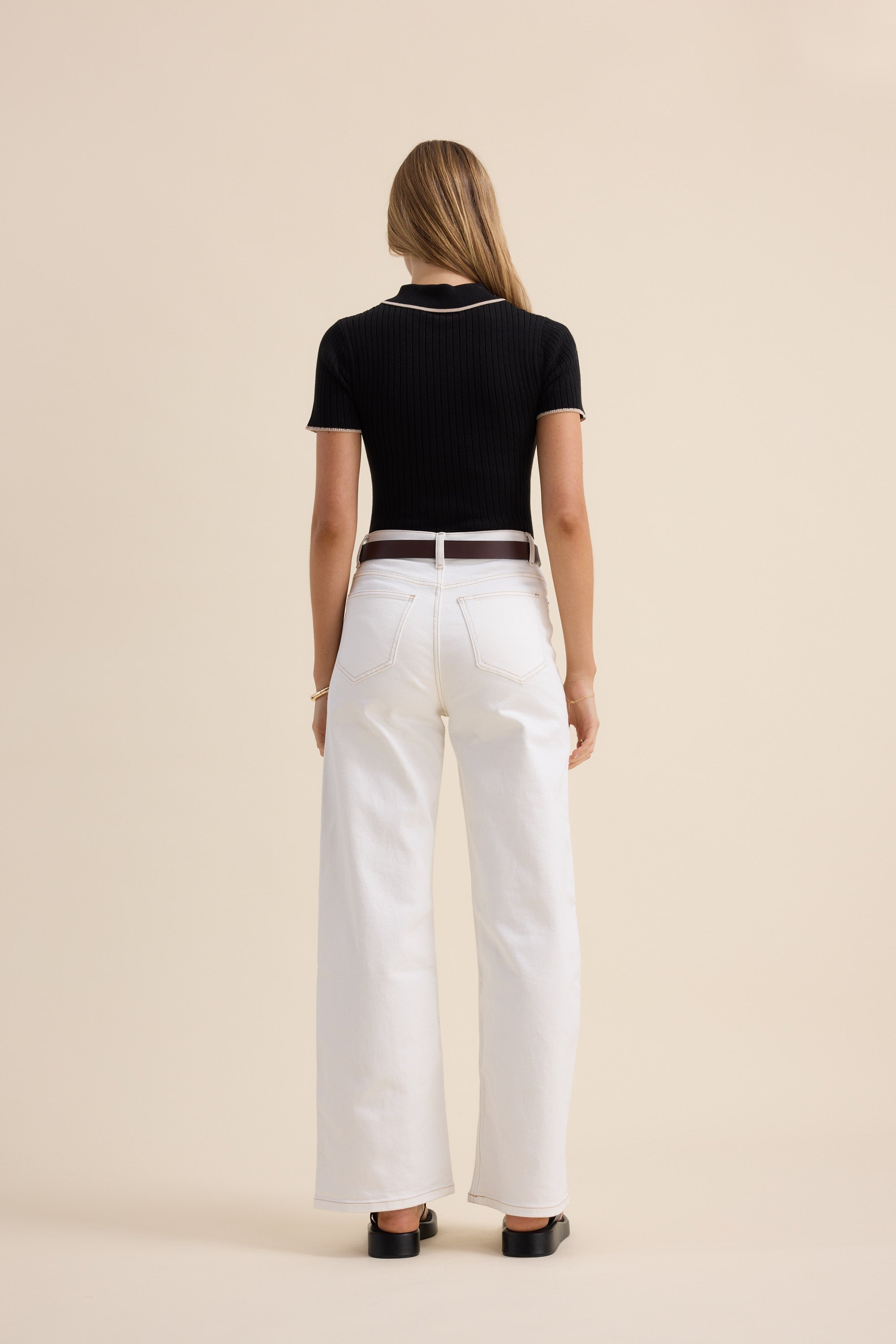 Woman wears Max’s The Knitted Henley Top in black, paired with white pants, back to camera.