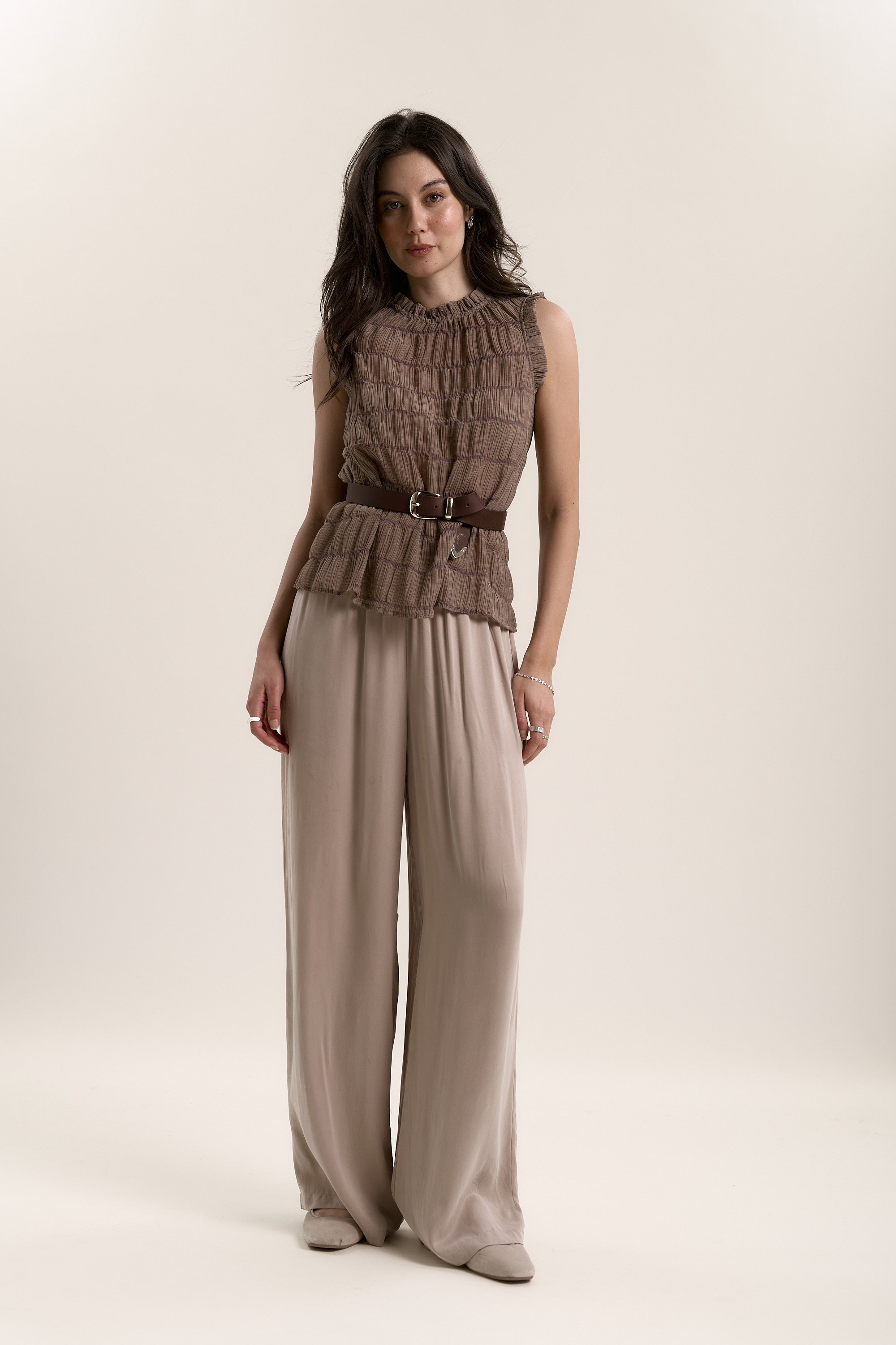 A woman with long dark hair and a neutral expression wears the Max Mindy Textured Top—sleeveless with ruffle details and a belt—paired with wide-legged beige pants, standing against a plain background.