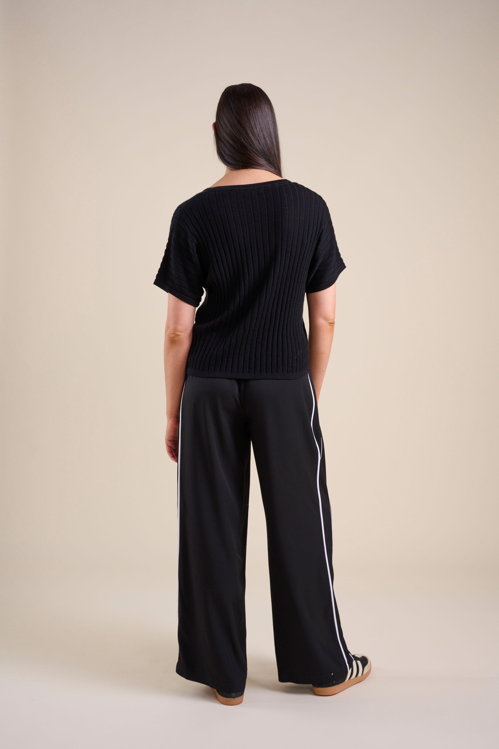 A woman faces away, wearing Max's The Elevated Jogger—loose striped pants with white side stripes—and a black short-sleeve ribbed top, set against a plain beige background.