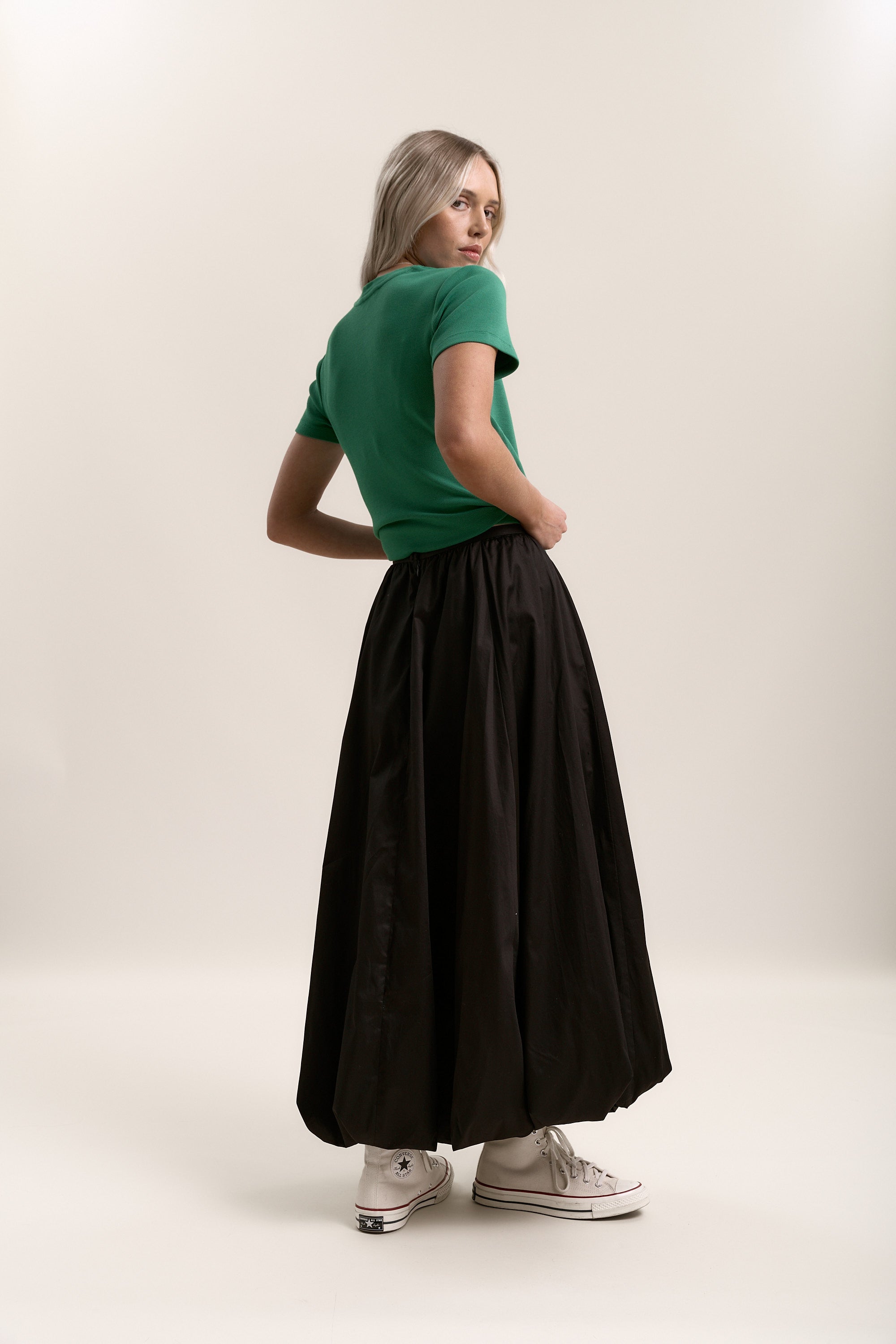 Woman in green top, Max Juniper Bubble Hem Skirt, and white sneakers poses by plain background.