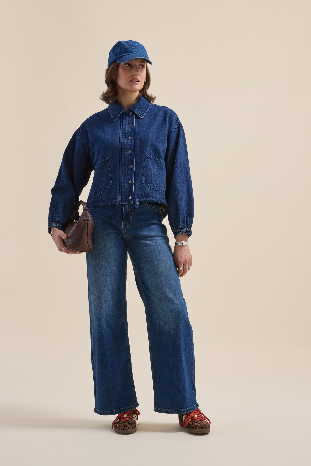 Model in Elementary Relaxed Jacket, jeans, blue cap, brown bag, red sandals on plain backdrop.