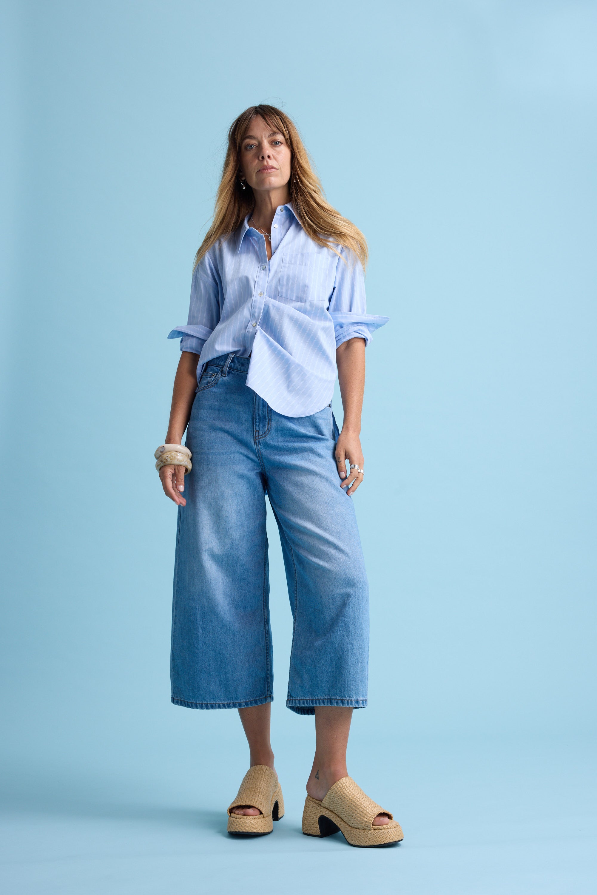 A woman stands against a light blue background, wearing Max Derby Wide Leg Cropped Jeans, a loose blue striped shirt, beige platform sandals, and several bracelets. Her long hair is loose and her hands rest at her sides.